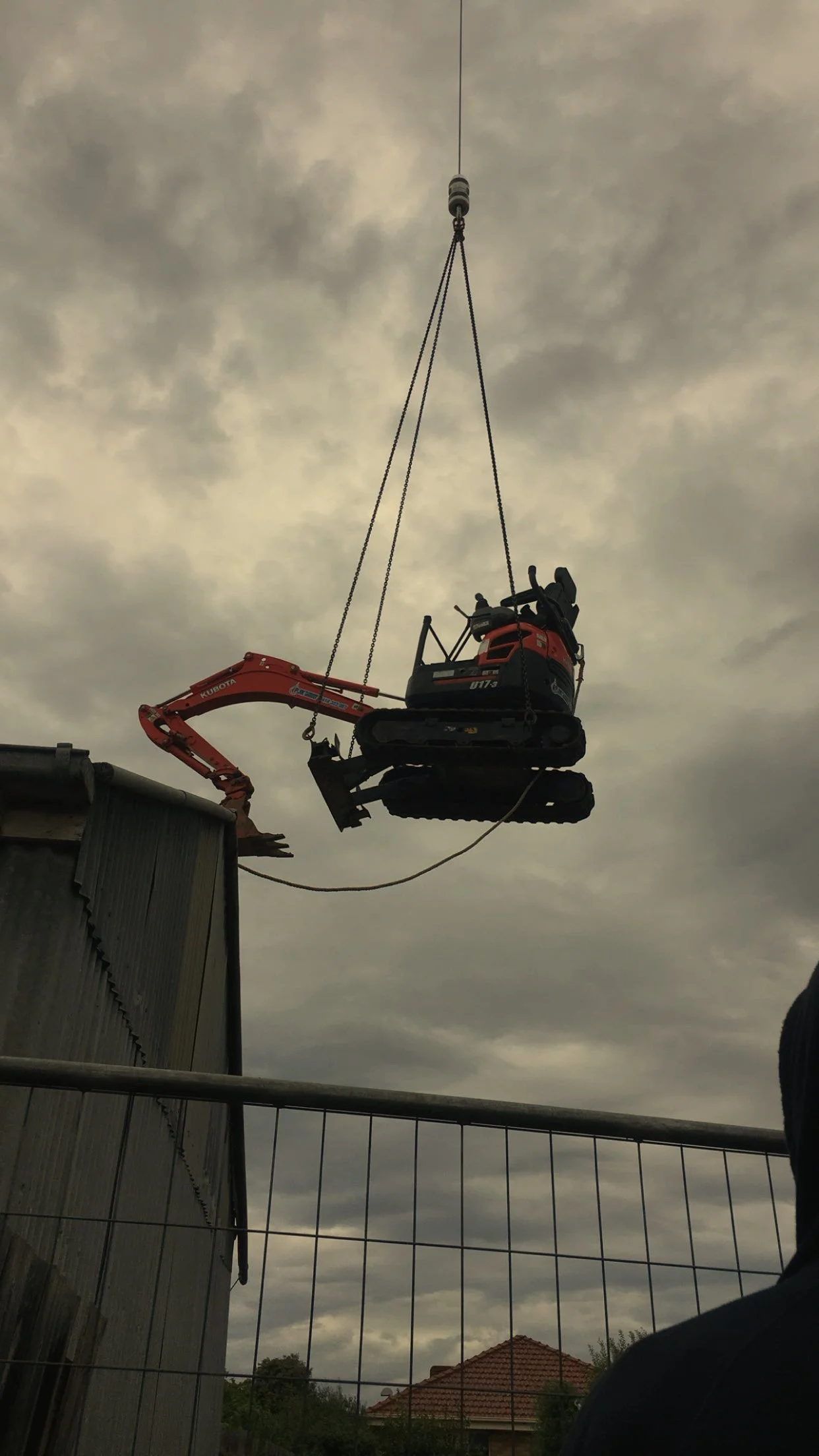 A small excavator being hoisted by a cable onto the roof of a building, with a cloudy sky in the background.
