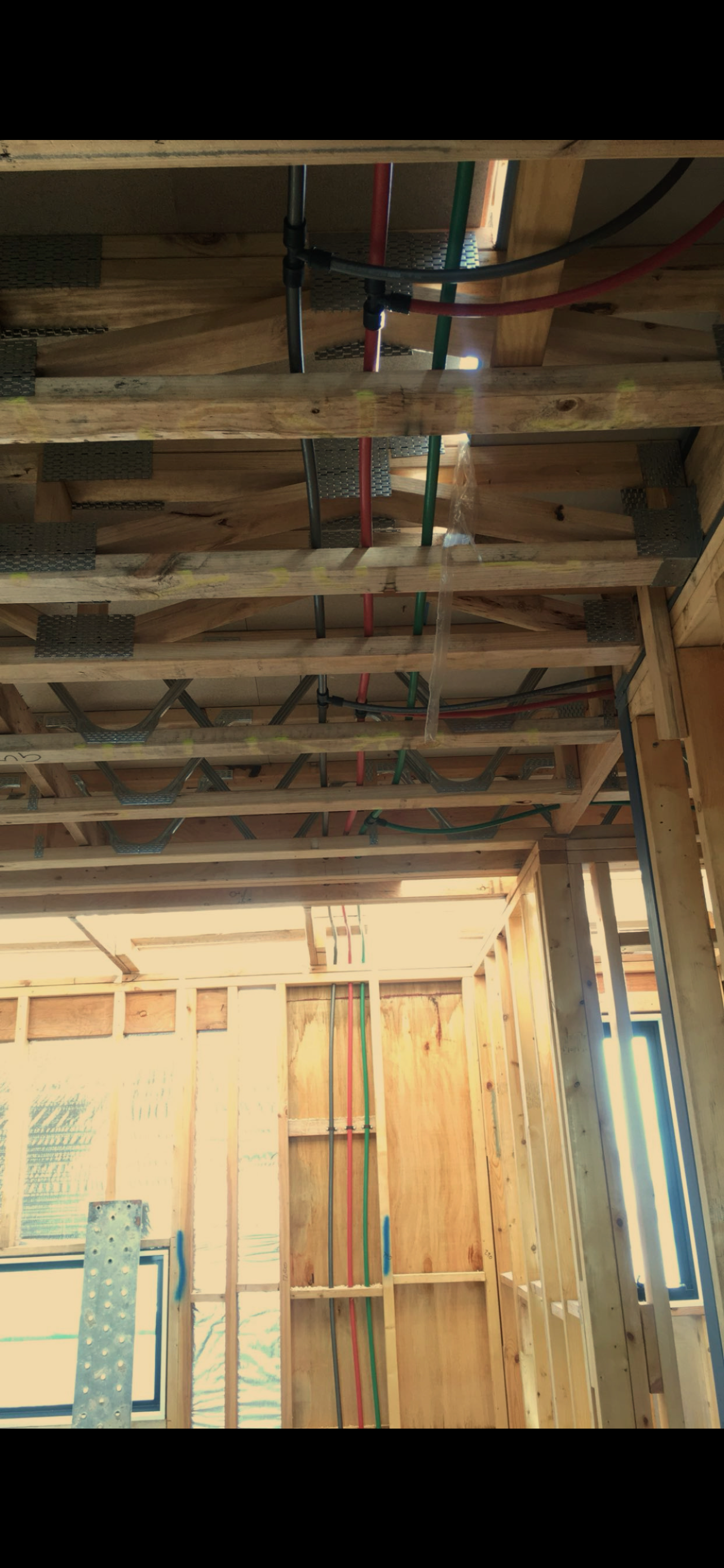 Interior view of a building under construction showing exposed wooden framing, electrical conduit, and plastic piping installed within the ceiling and walls.