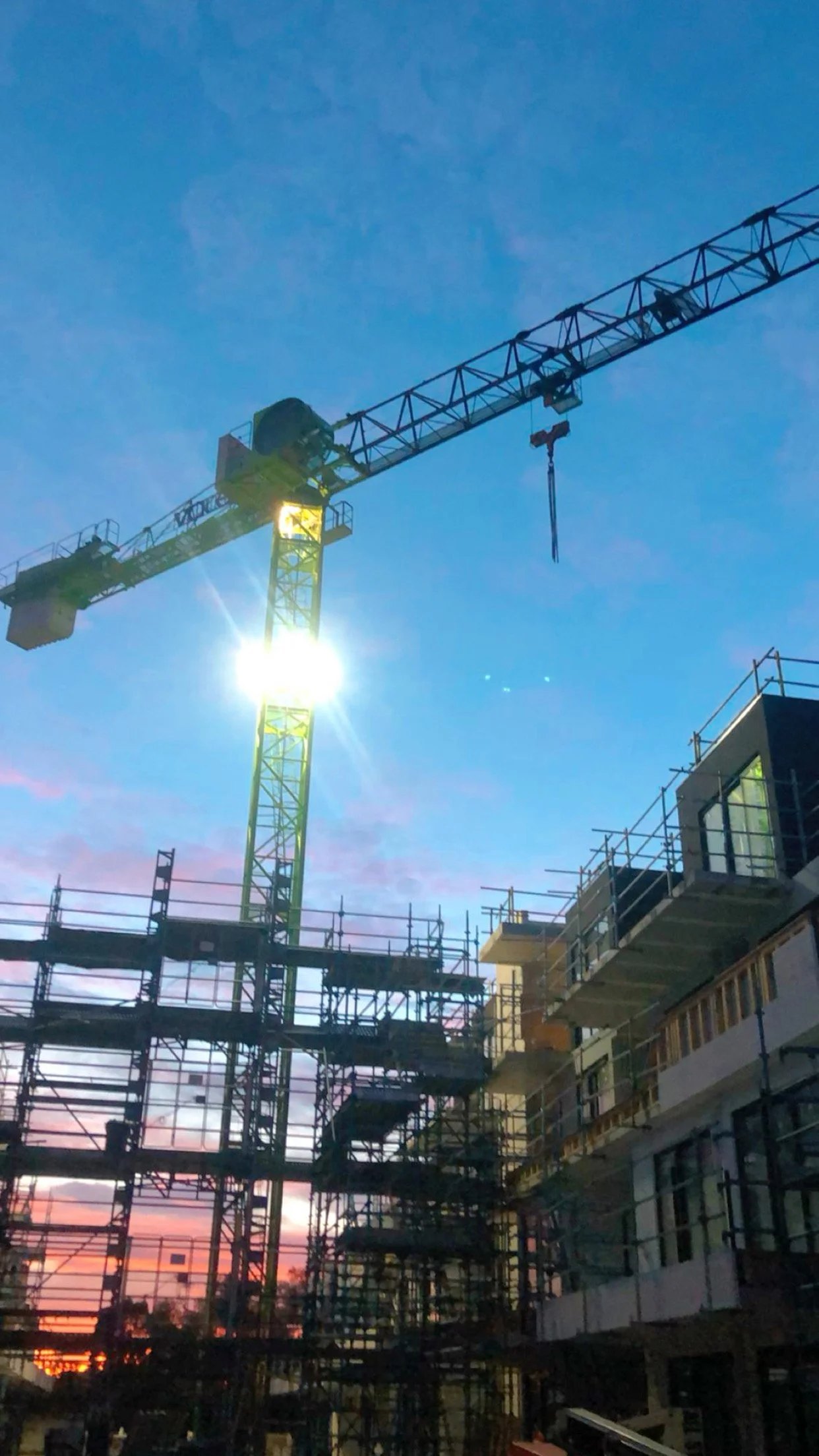 Construction site with a tall tower crane against a blue sky at sunset, scaffolding and partially built buildings in the foreground.
