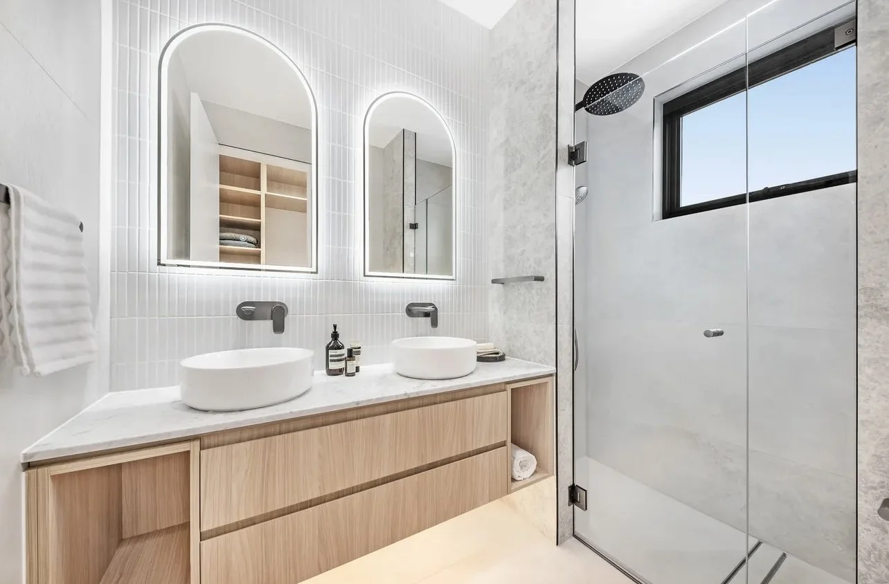 Modern bathroom with double vanity sinks, illuminated mirrors, a glass shower enclosure, a window, and minimalist decor.