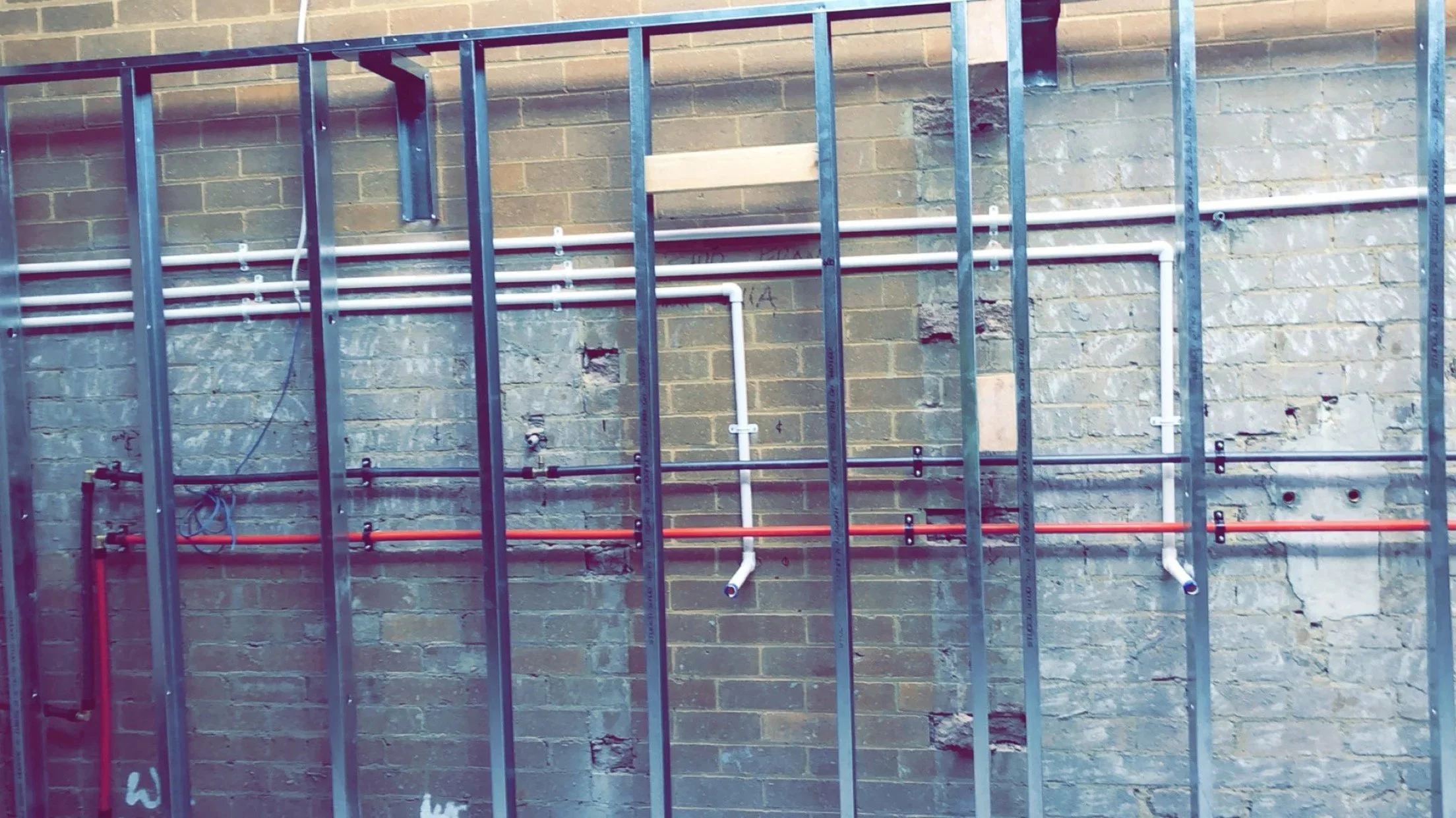 Construction wall with metal framing and exposed pipes in various colors against a brick wall.