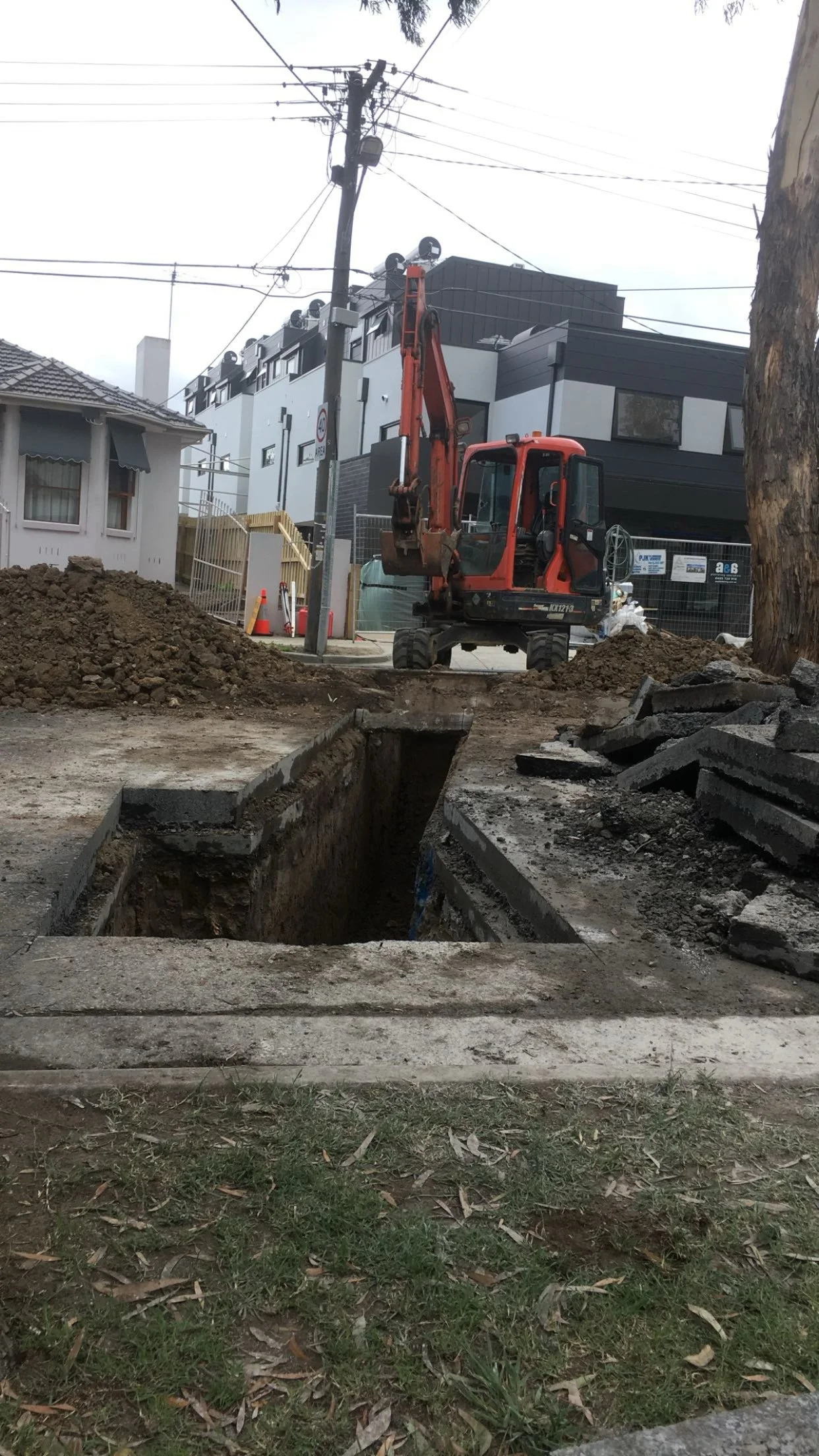 Construction site with an excavator digging a trench on a city street, surrounded by soil and construction materials, with buildings and utility poles in the background.