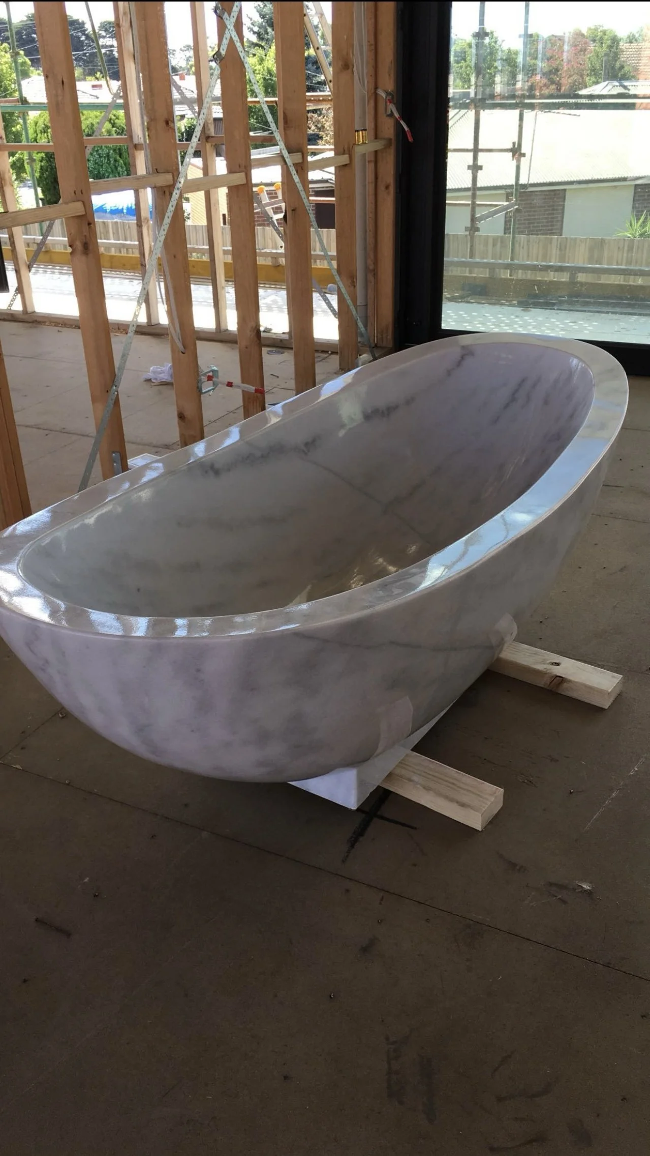 Marble bathtub on wooden supports in a construction site with exposed wooden framing and large glass windows.