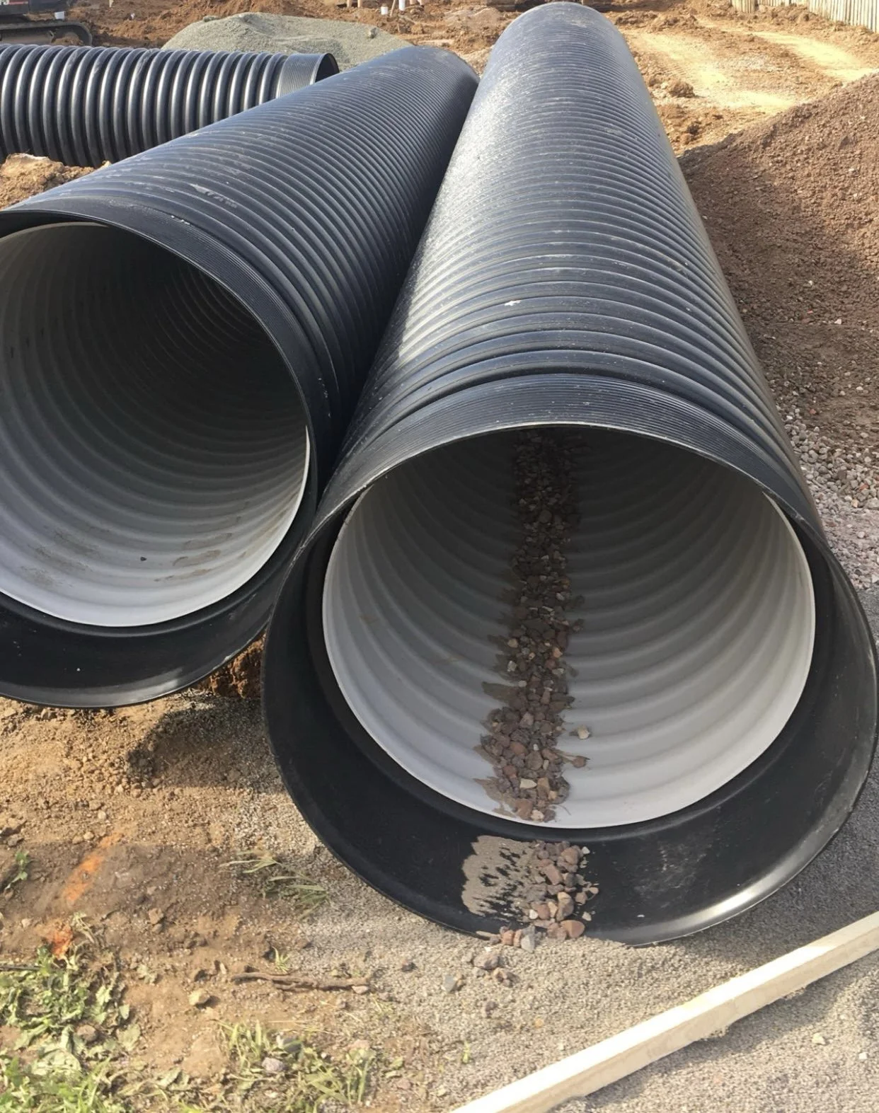 Large black and white corrugated plastic pipes lying on a construction site with gravel and dirt ground.