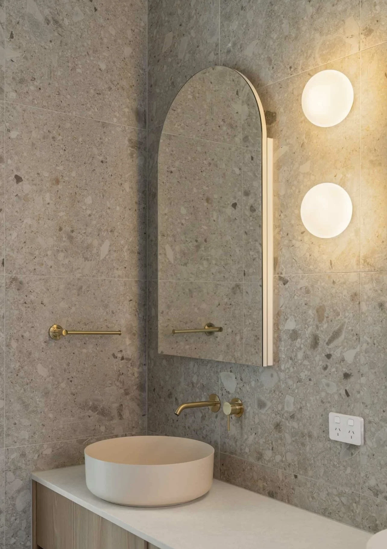 Modern bathroom with beige textured wall tiles, a round vessel sink, gold fixtures, an arched mirror, two globe wall lights, and a white electrical outlet.