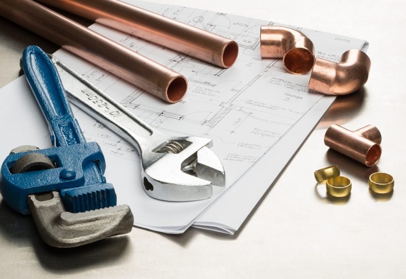 Plumbing tools and materials, including copper pipes, a pipe wrench, pipe fittings, and plumbing blueprints.