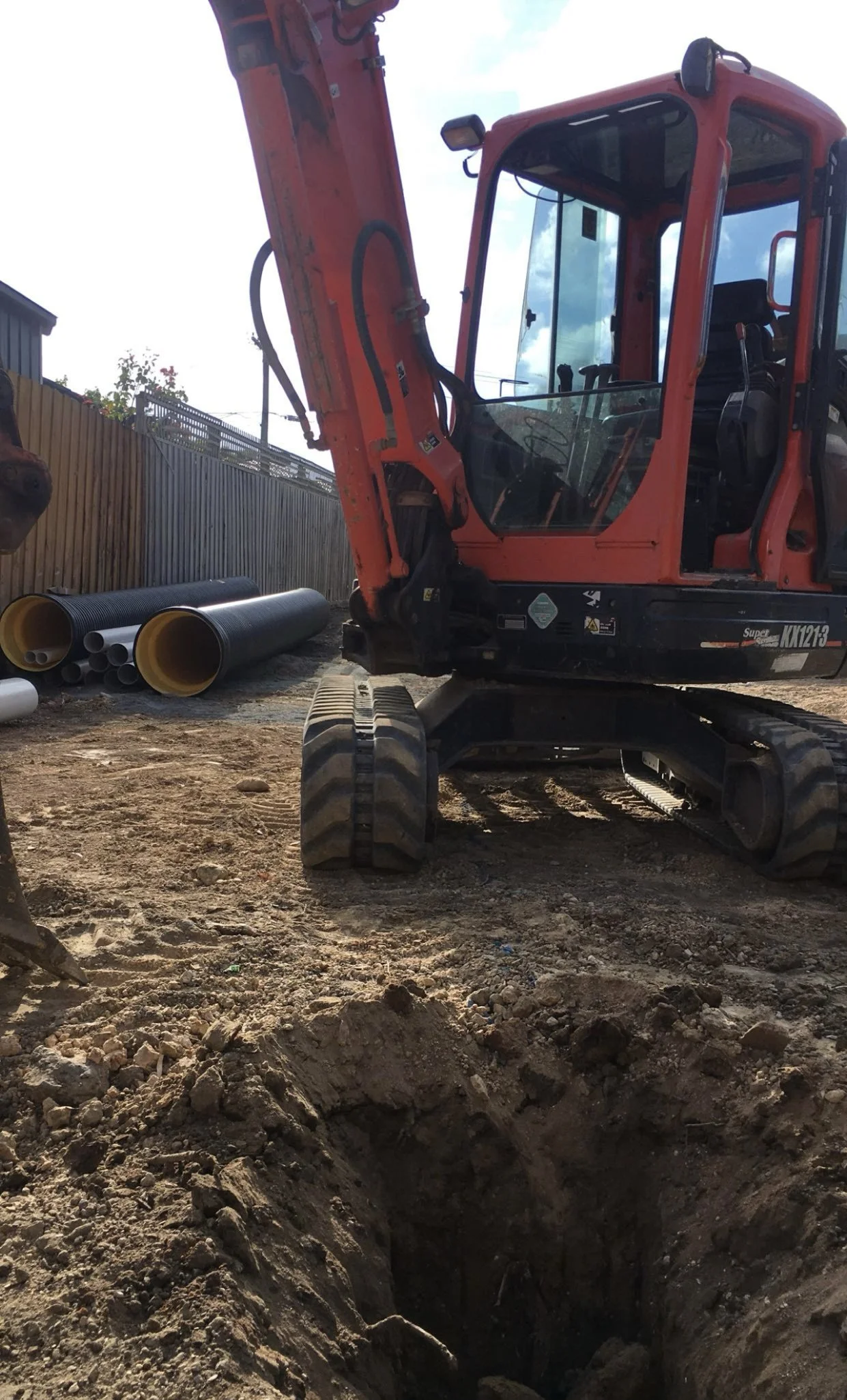 Construction site with a small orange excavator, dirt in a trench, and pipes stacked nearby.
