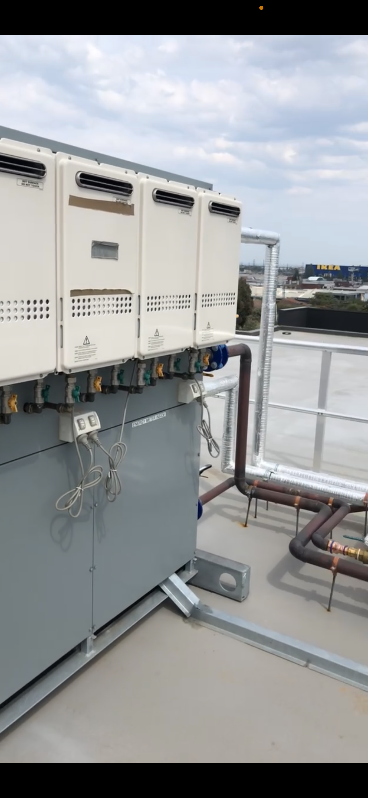 Rooftop HVAC unit with multiple vented metal boxes, thermostats, and associated piping against a cloudy sky, with a distant view of a commercial area including an IKEA store.