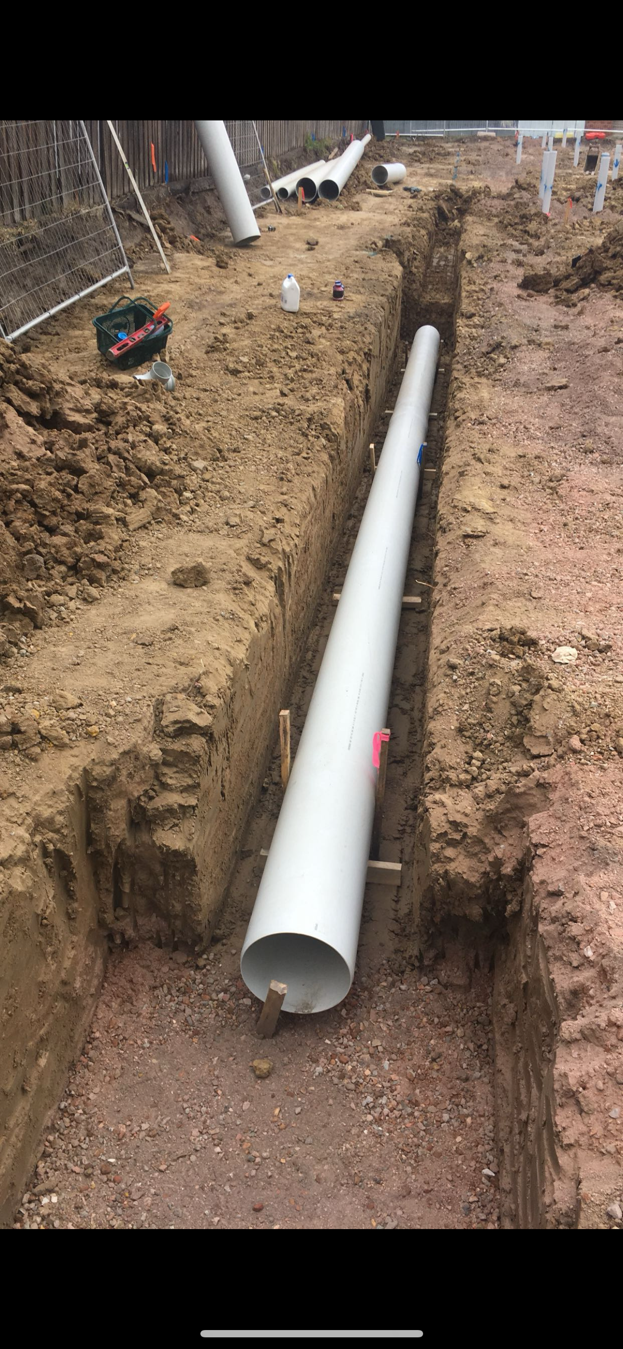 Construction site with large white pipe laid in a long trench, surrounded by dirt and construction tools.