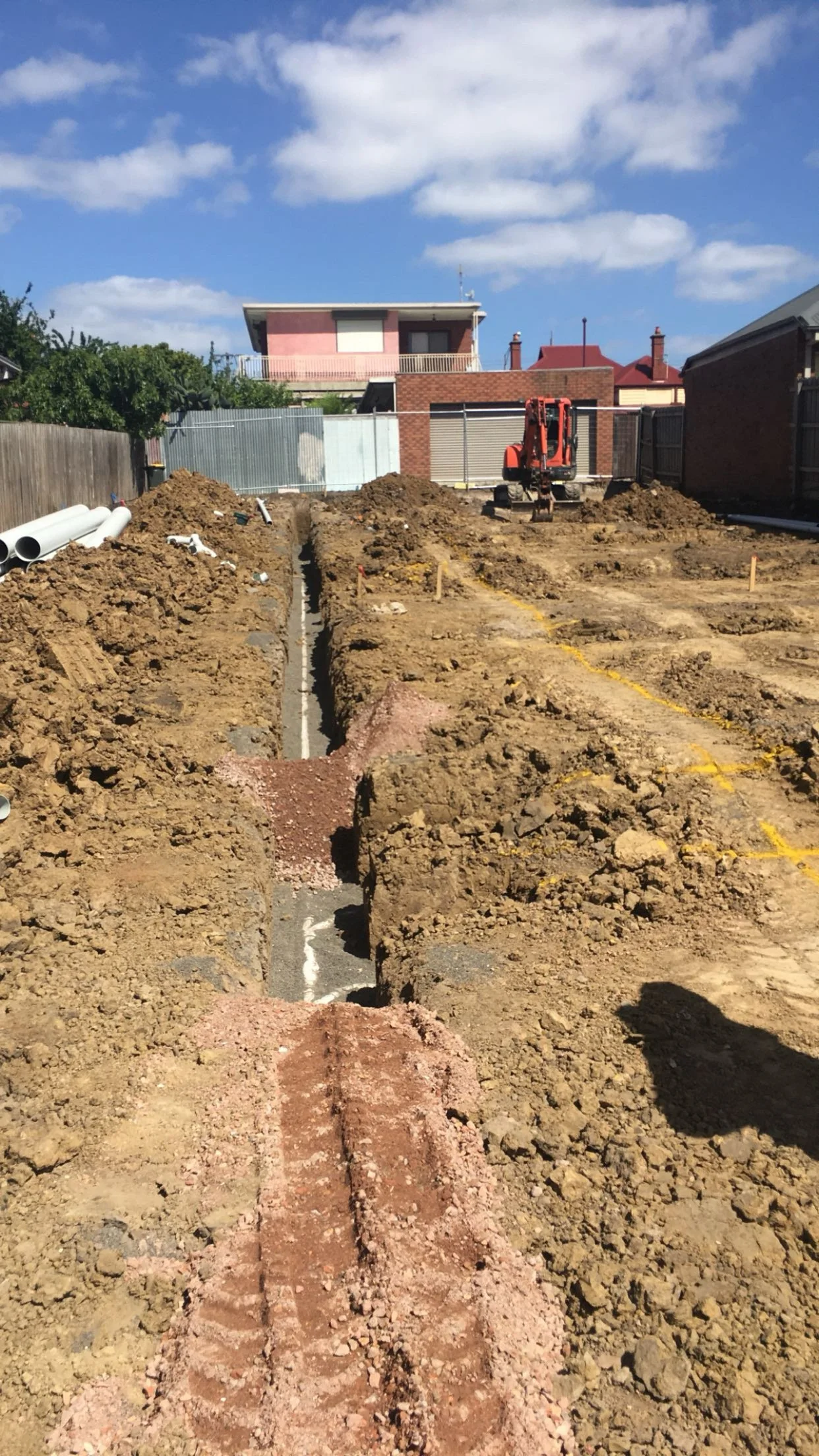 Construction site with dirt trenches and a small excavator in a backyard under a partly cloudy sky.