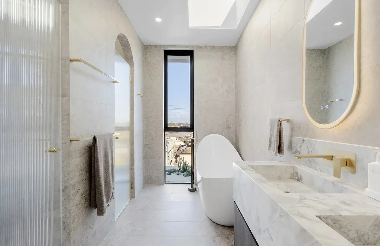 Modern bathroom with a freestanding bathtub, marble countertop sink, large mirror, tall window, and beige walls.