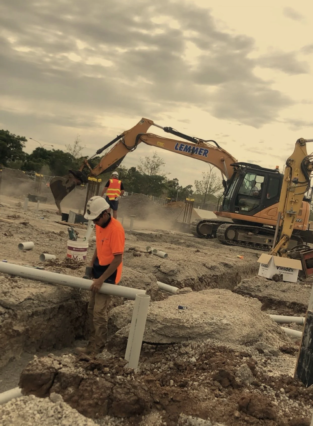 Workers at a construction site installing pipes, with an excavator in the background and overcast sky above.