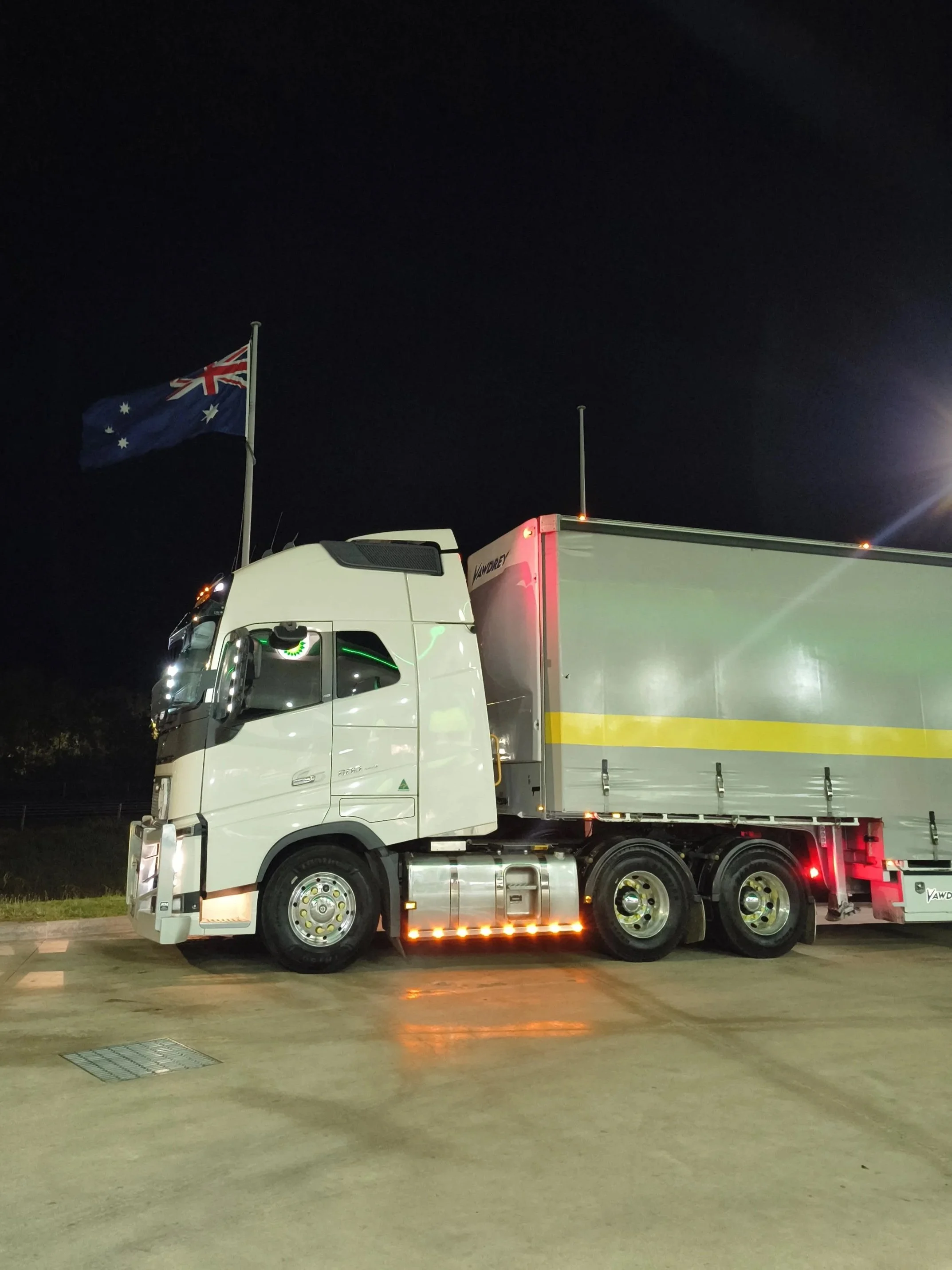 Nighttime view of a large white semi-truck with a gray trailer, illuminated by lights, parked on a concrete surface underneath a dark sky. An Australian flag is flying on a flagpole next to the truck.