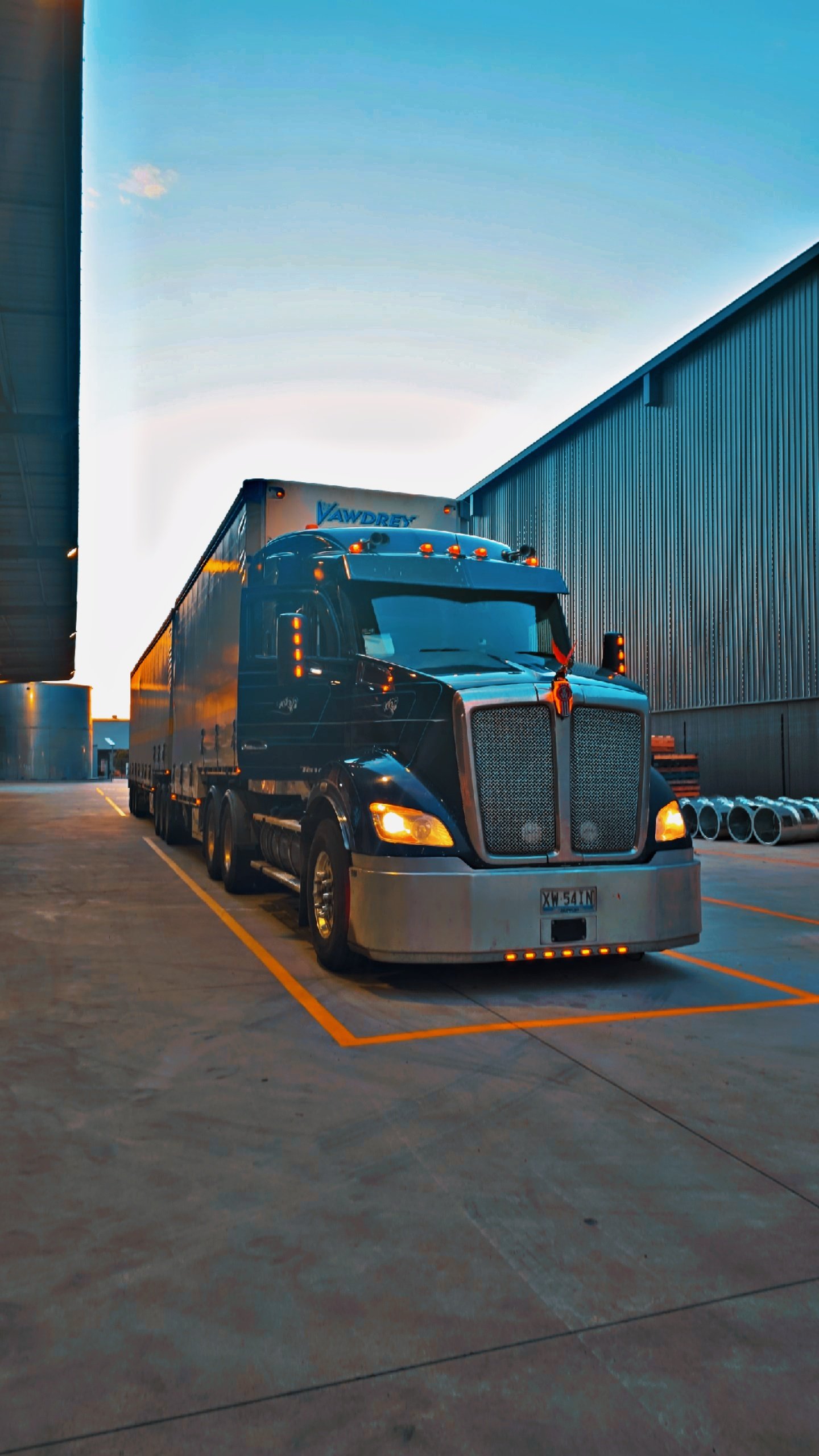 A large black semi-truck with orange lights parked in a loading dock with industrial buildings on either side, showing a clear evening sky.
