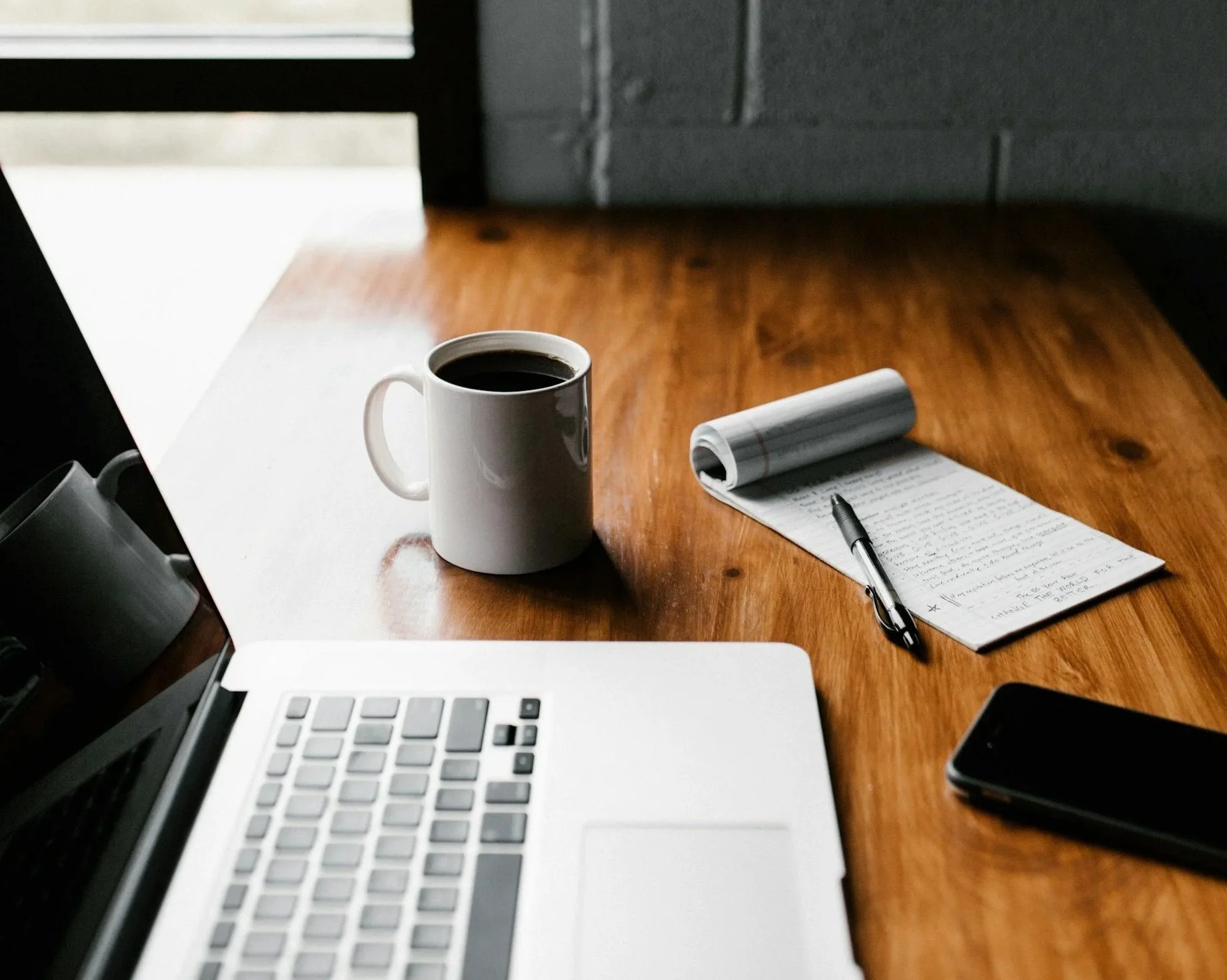 A wooden desk with a laptop, a cup of coffee, a notepad with handwritten notes, a pen, and a smartphone.