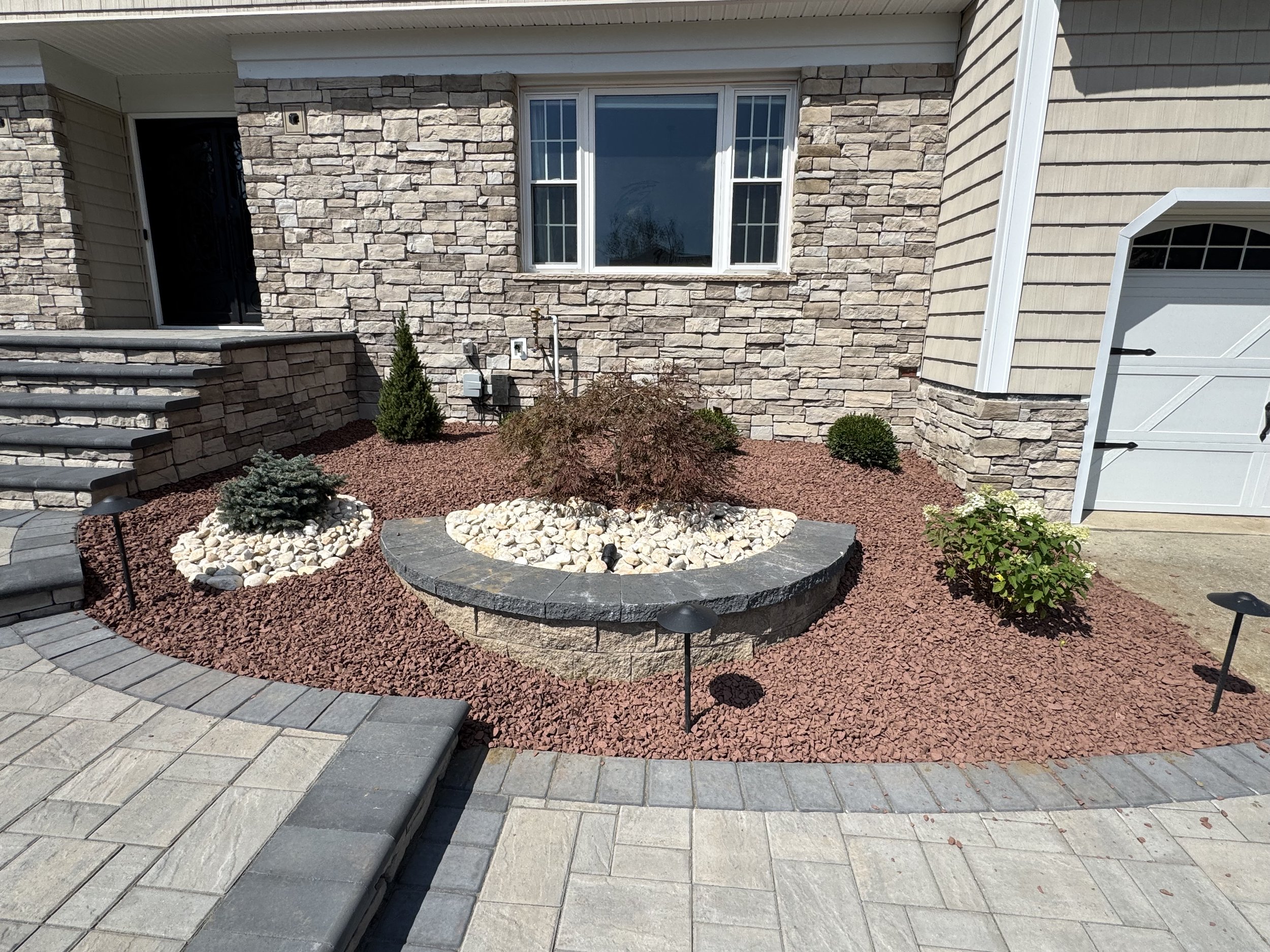 Front yard with stone landscaping and small shrubs, paved walkway, steps, and a brick house with window and garage door.