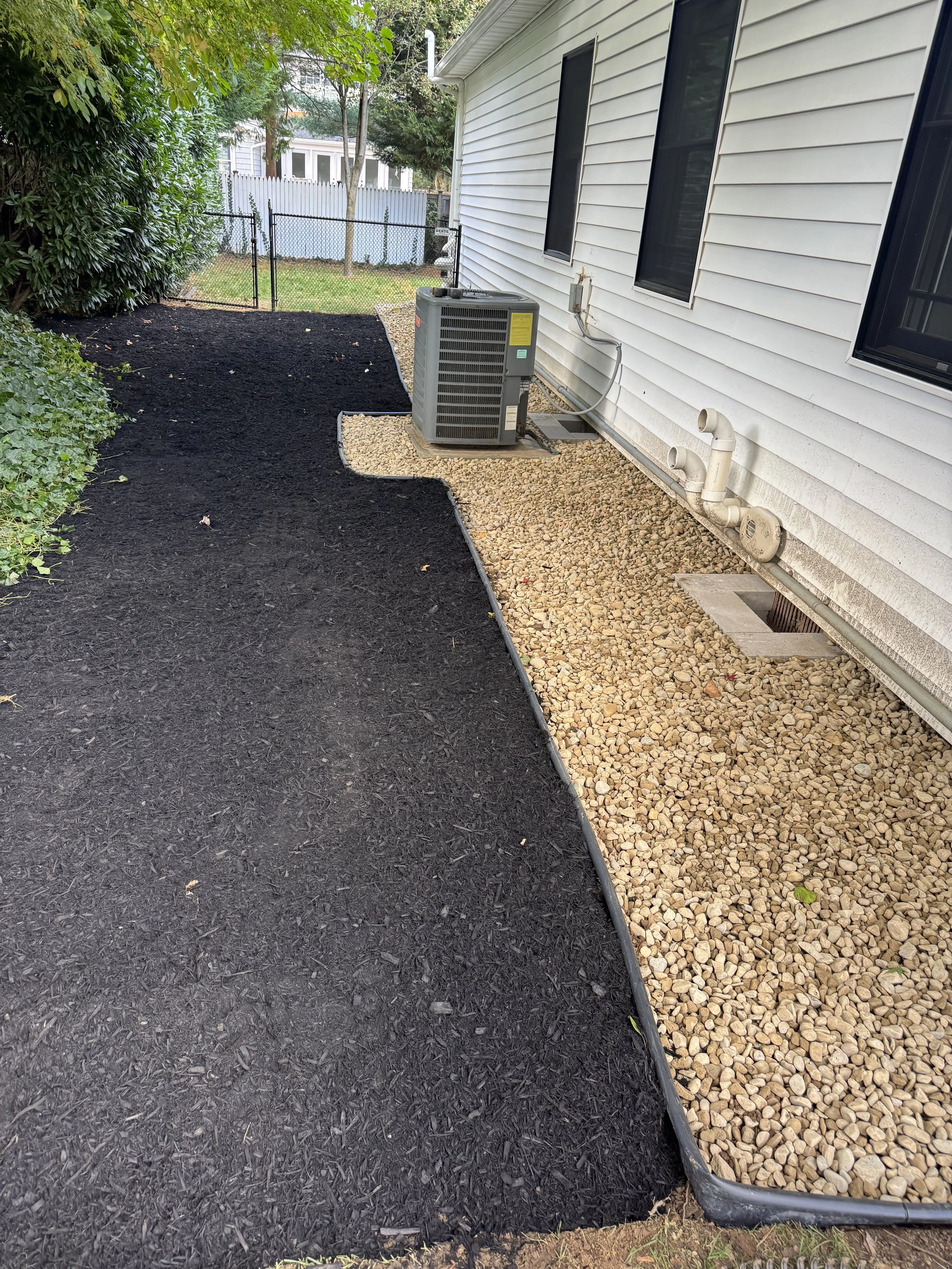 A backyard with a black mulch area, a gravel pathway, and a white house with a visible HVAC unit on the ground.
