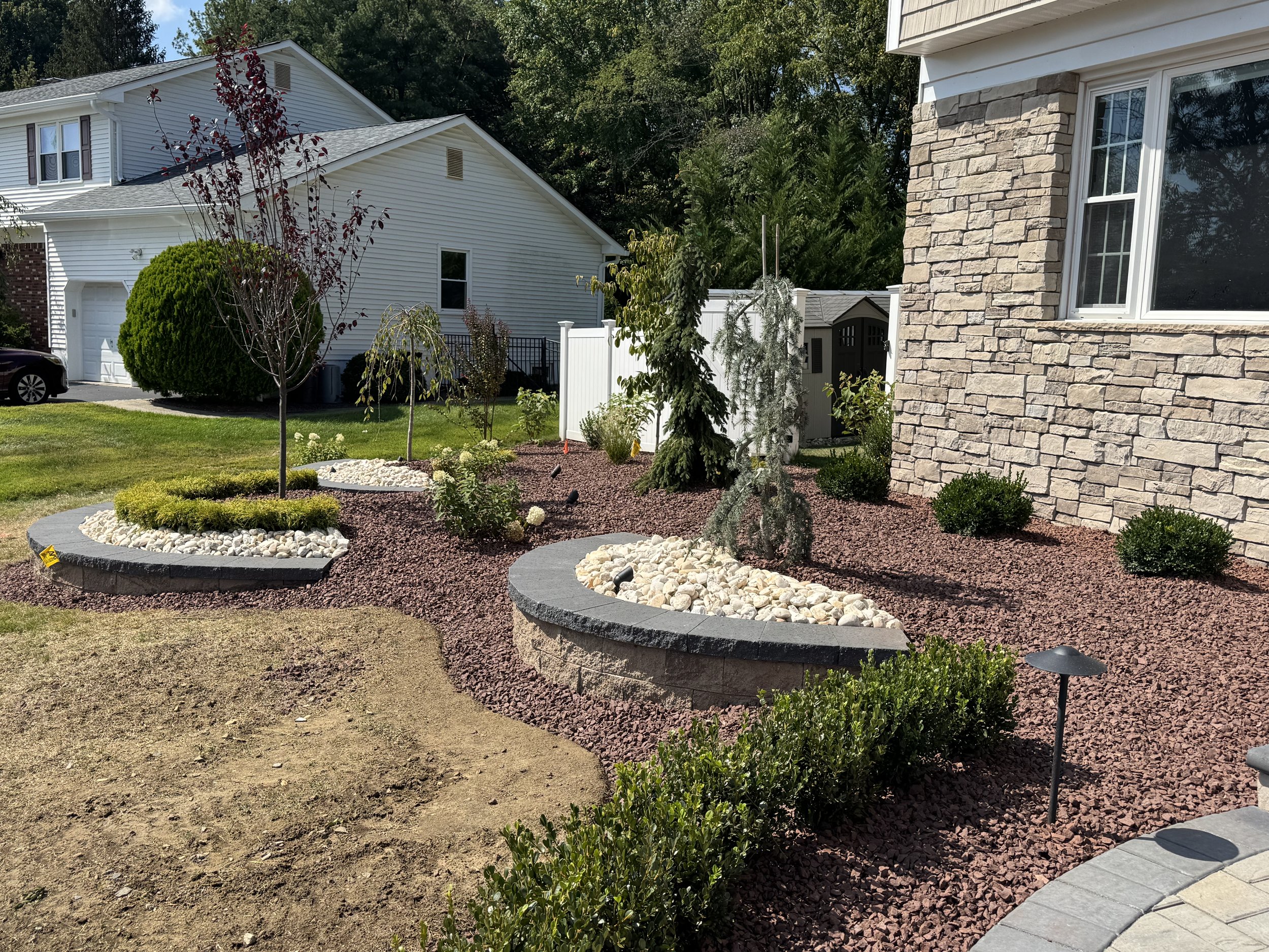 A landscaped front yard with trees, bushes, and decorative rocks surrounded by mulch, next to a house with a stone exterior and a large window.
