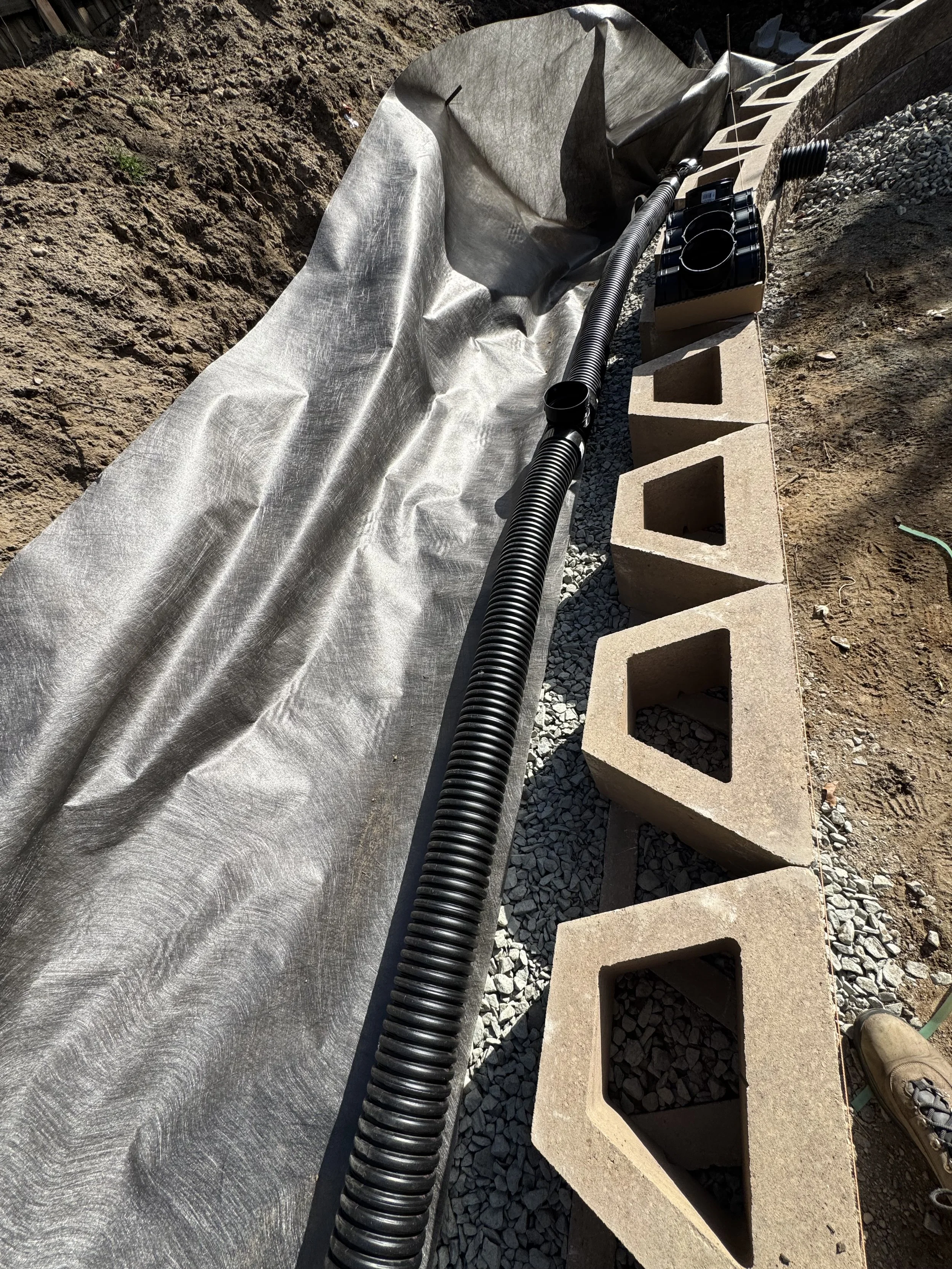Construction site with black corrugated pipe, gray fabric liner, concrete cinder blocks with a french drain being installed into the wall.