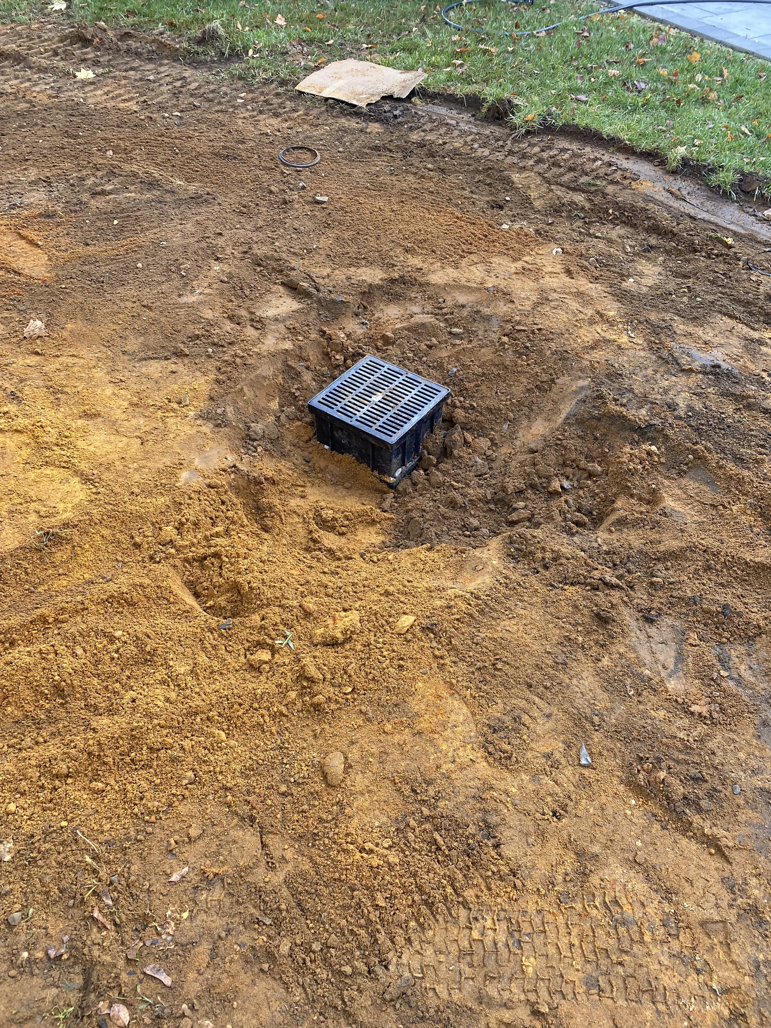A small black drainage grate surrounded by dirt and soil in an outdoor area with grass and construction material nearby.