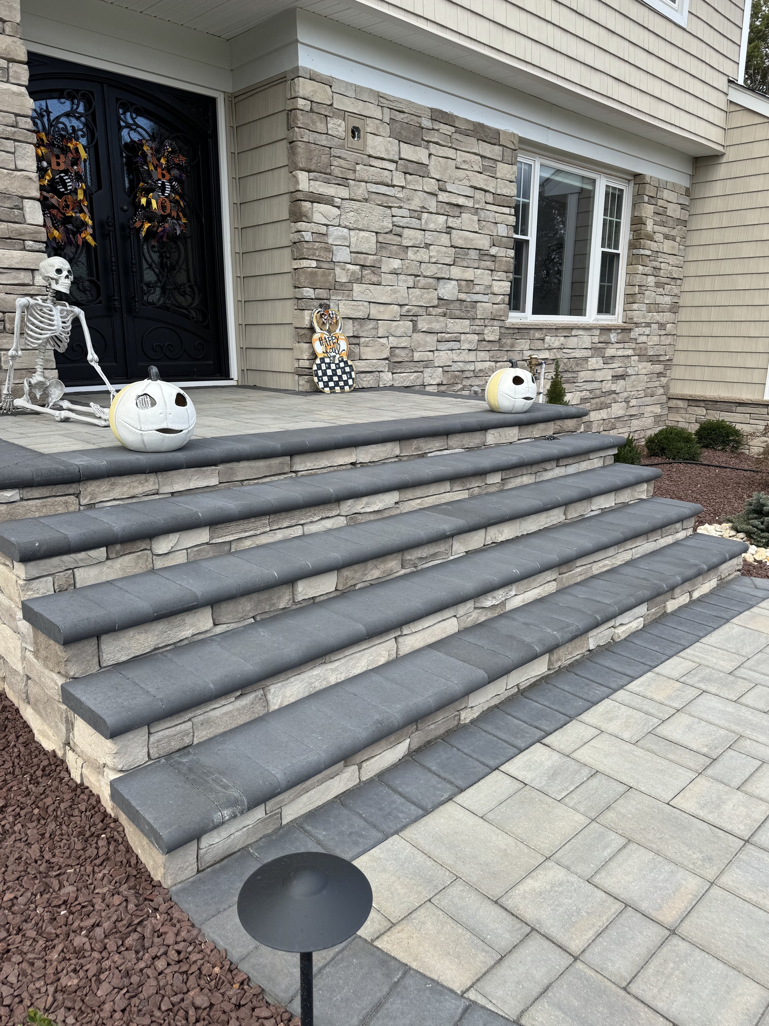 Front porch decorated for Halloween with skeleton and pumpkin decorations on stone steps, including a black door with Halloween wreaths, and a stone veneer wall.