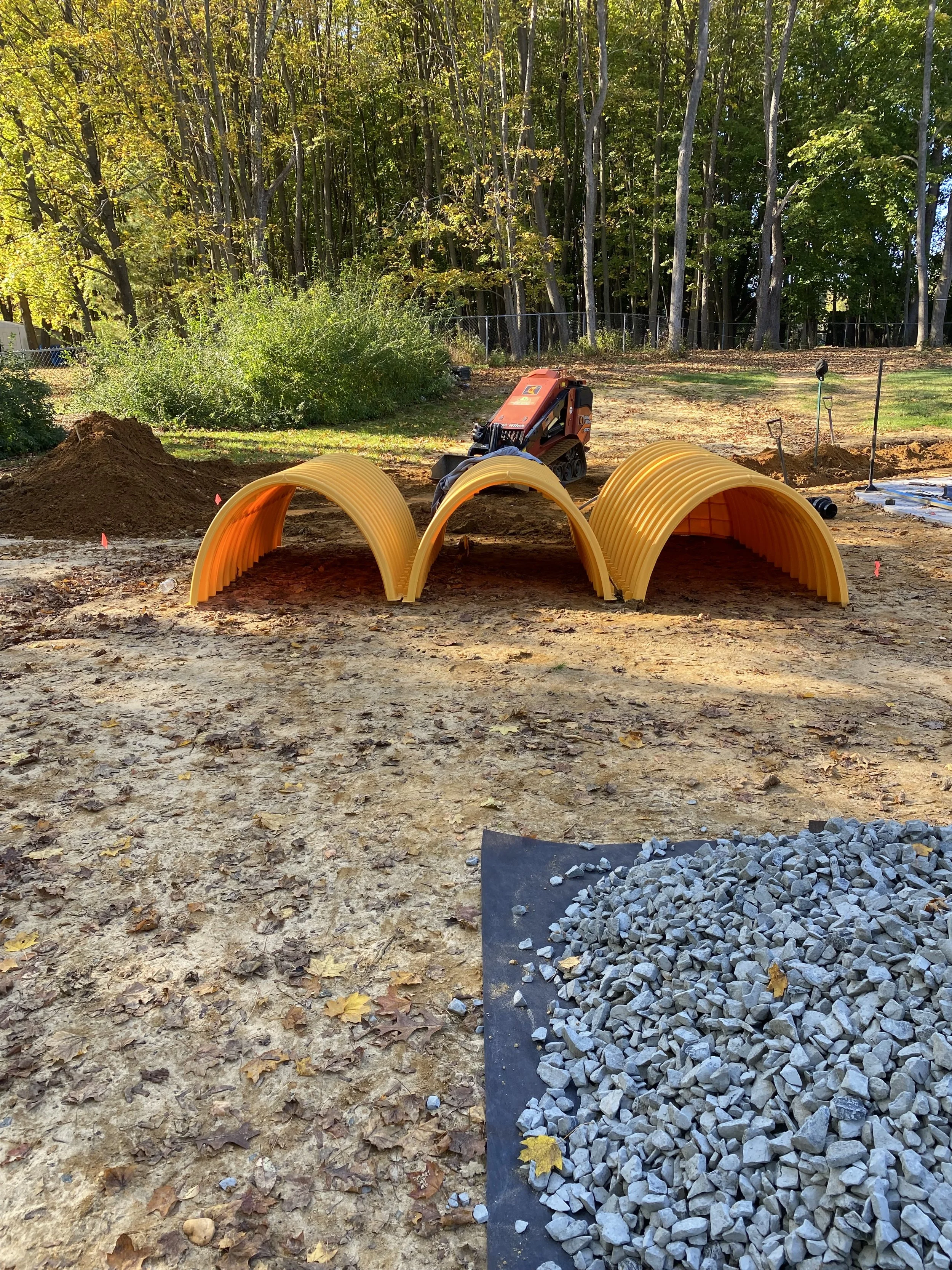 Construction site with orange curved pipes forming walkways, a small orange construction vehicle, gravel, dirt piles, and trees in the background.