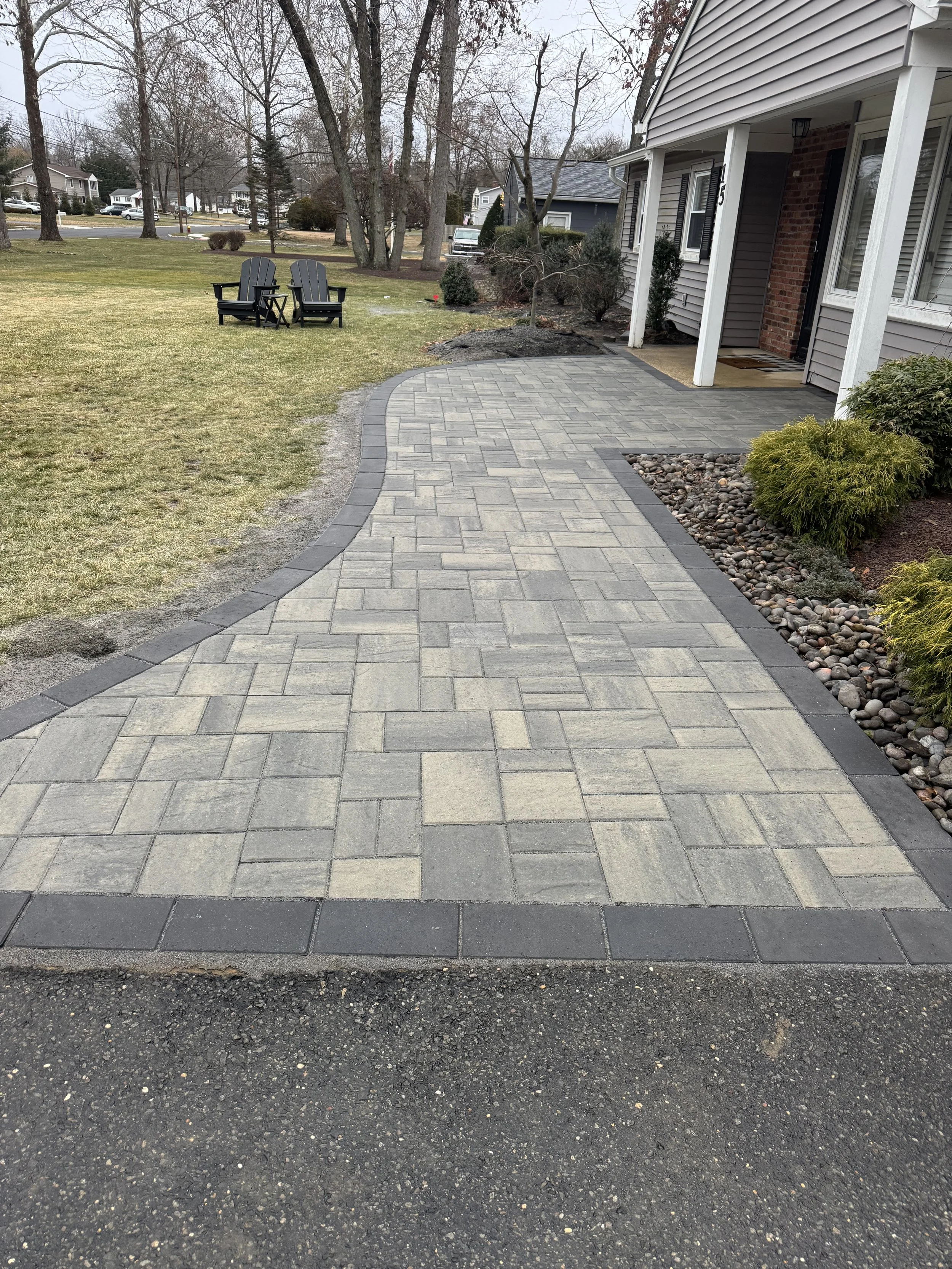 Paved walkway with a decorative border leading to a house, with a garden bed containing plants and rocks on one side, and a lawn with outdoor chairs in the background.