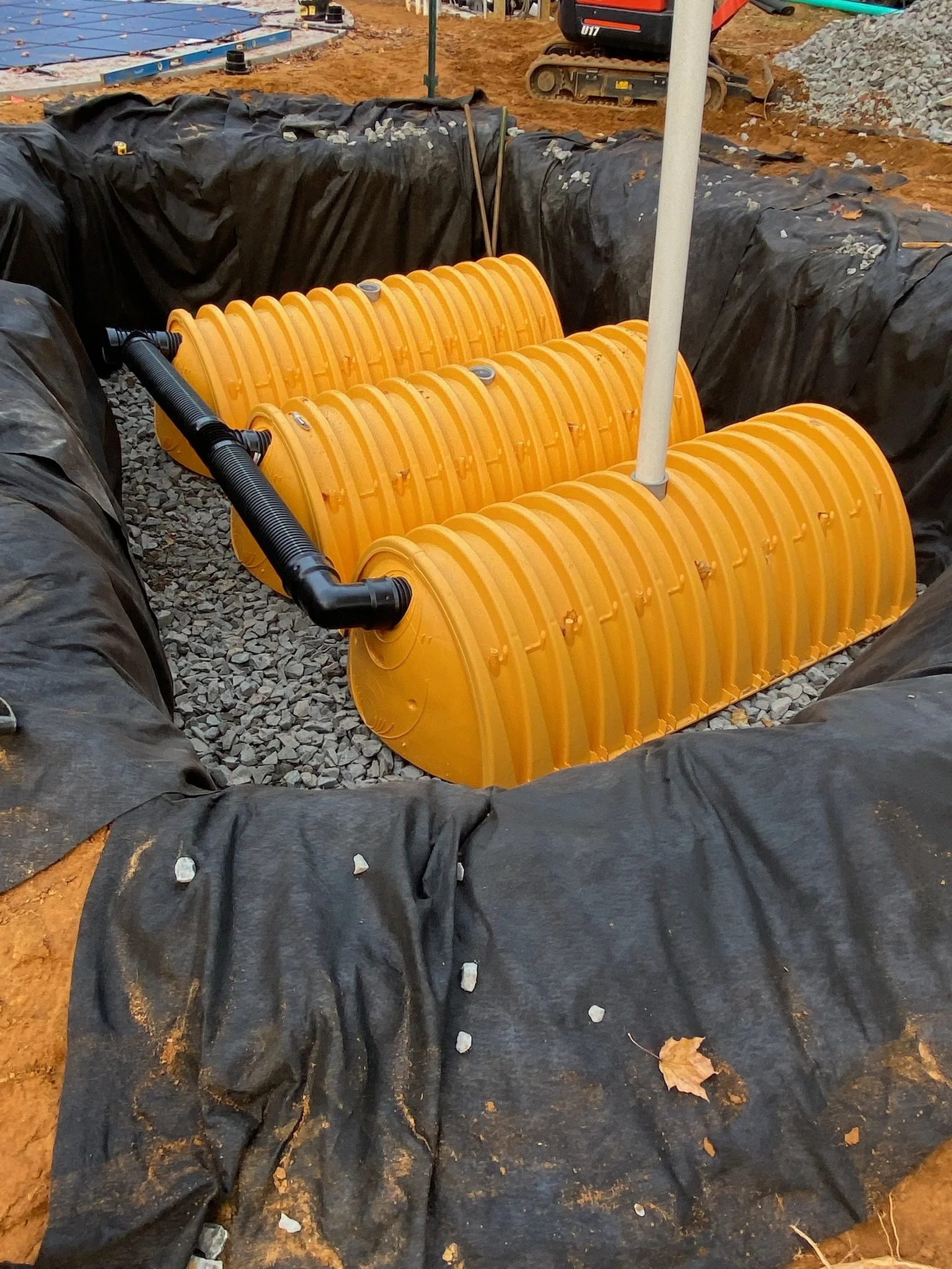 Underground utility vault with orange plastic covers, surrounded by black geotextile fabric and gravel, with pipes connecting to the covers at a construction site.