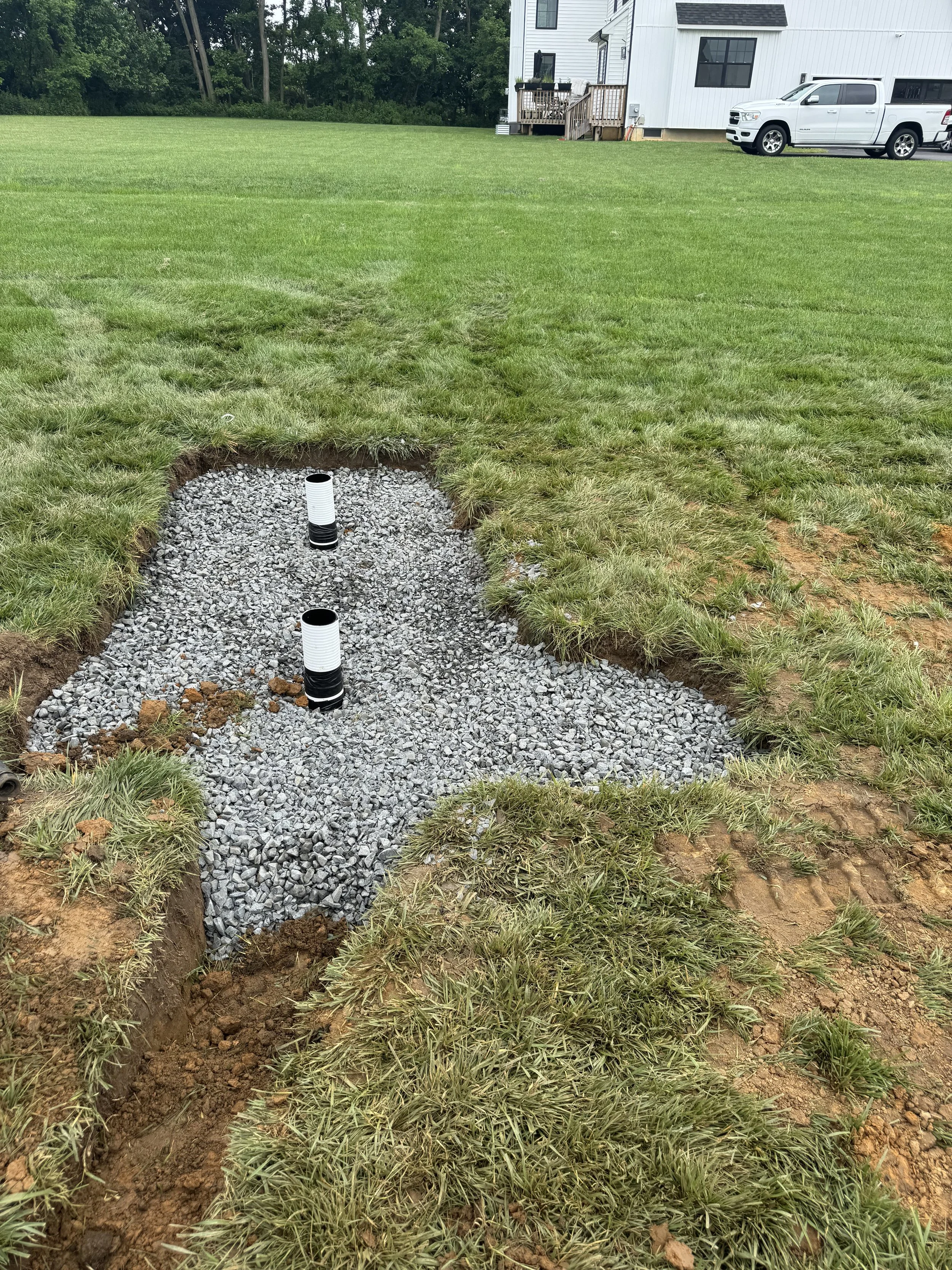 Two white and black pipes sticking out of a gravel trench in a grassy yard under construction, dry well being installed to hold excess water.