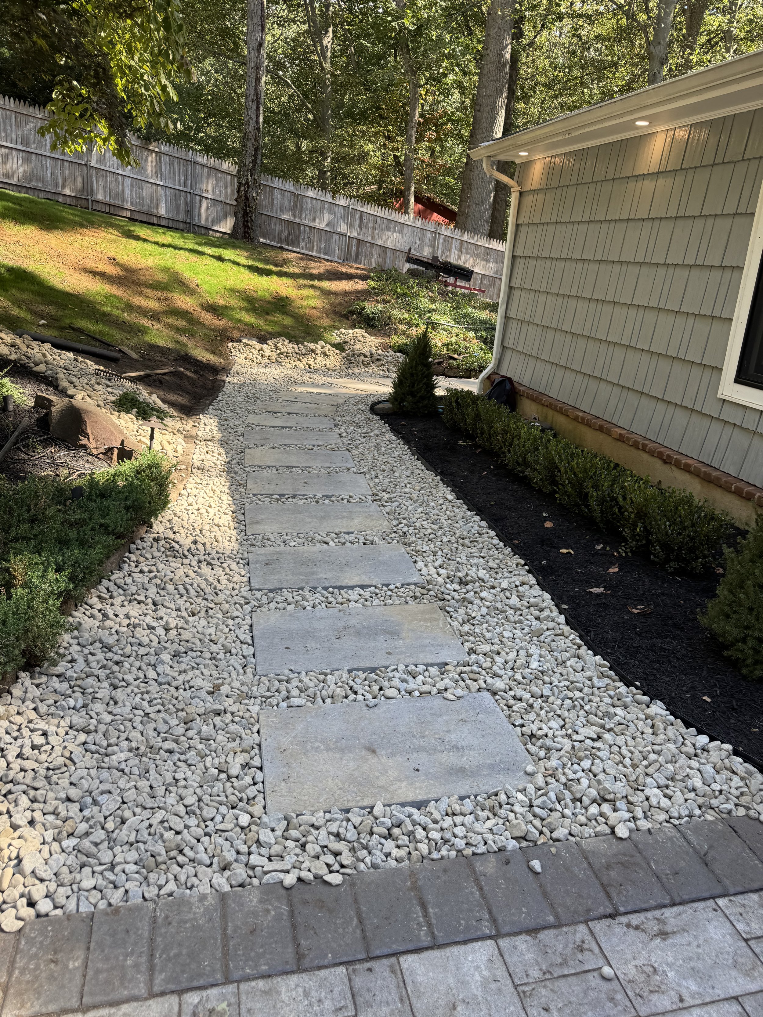 A landscaped backyard pathway made of large rectangular stepping stones surrounded by white gravel, bordered by green shrubs and a garden bed with dark mulch, with a wooden fence and trees in the background.
