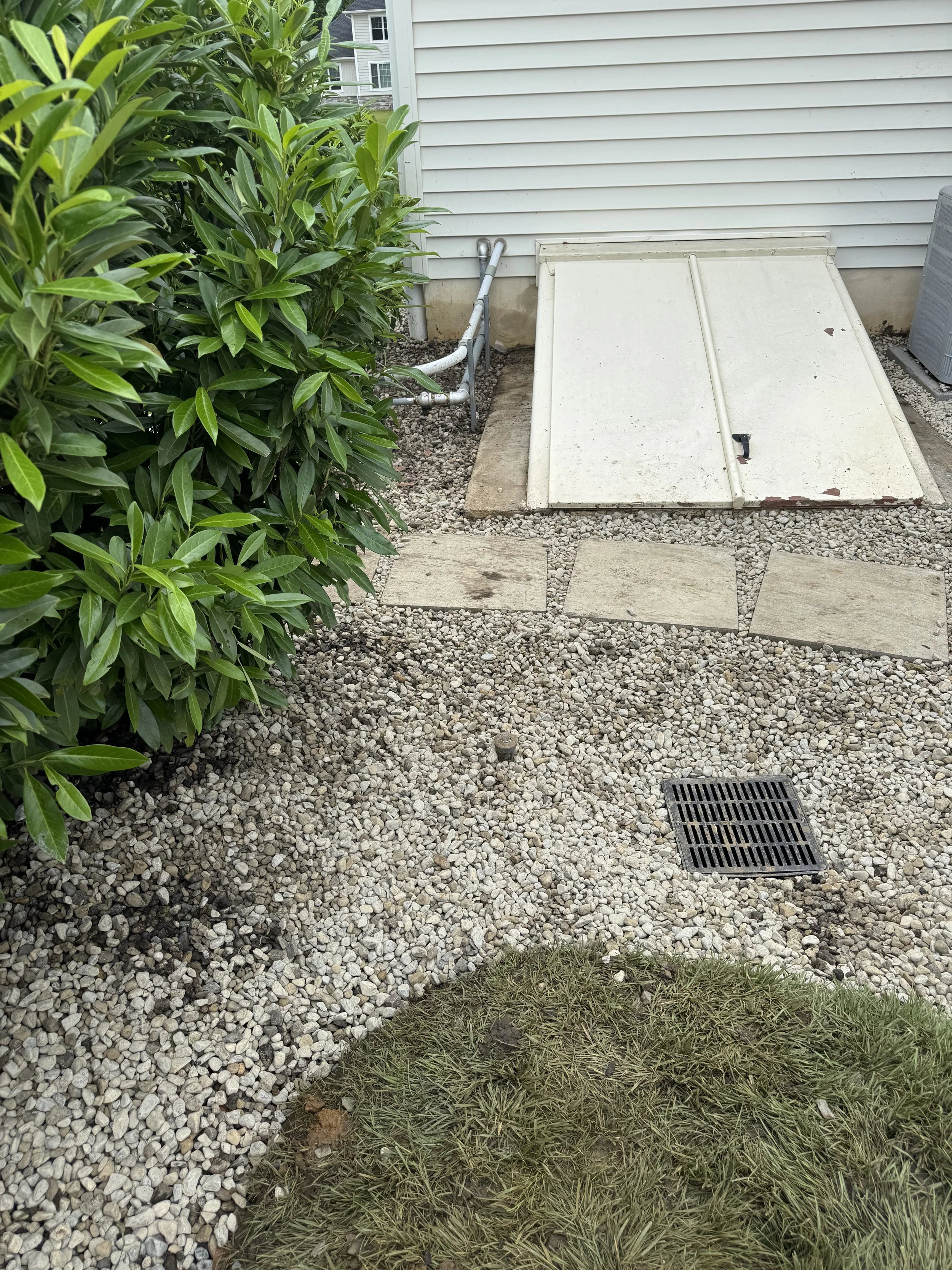 Side yard with gravel pathway, green bushes on the left, a white hatch door with a lock, a small concrete step, a drain grate, and a white house with siding in the background.
