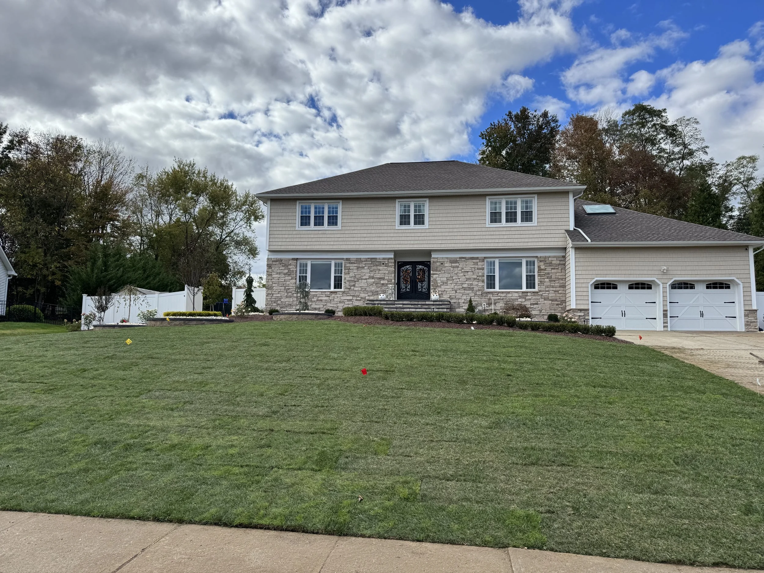 A large two-story house with a brown roof, beige siding, and a cultured stone facade. Sod to keeps water flowing away from house.