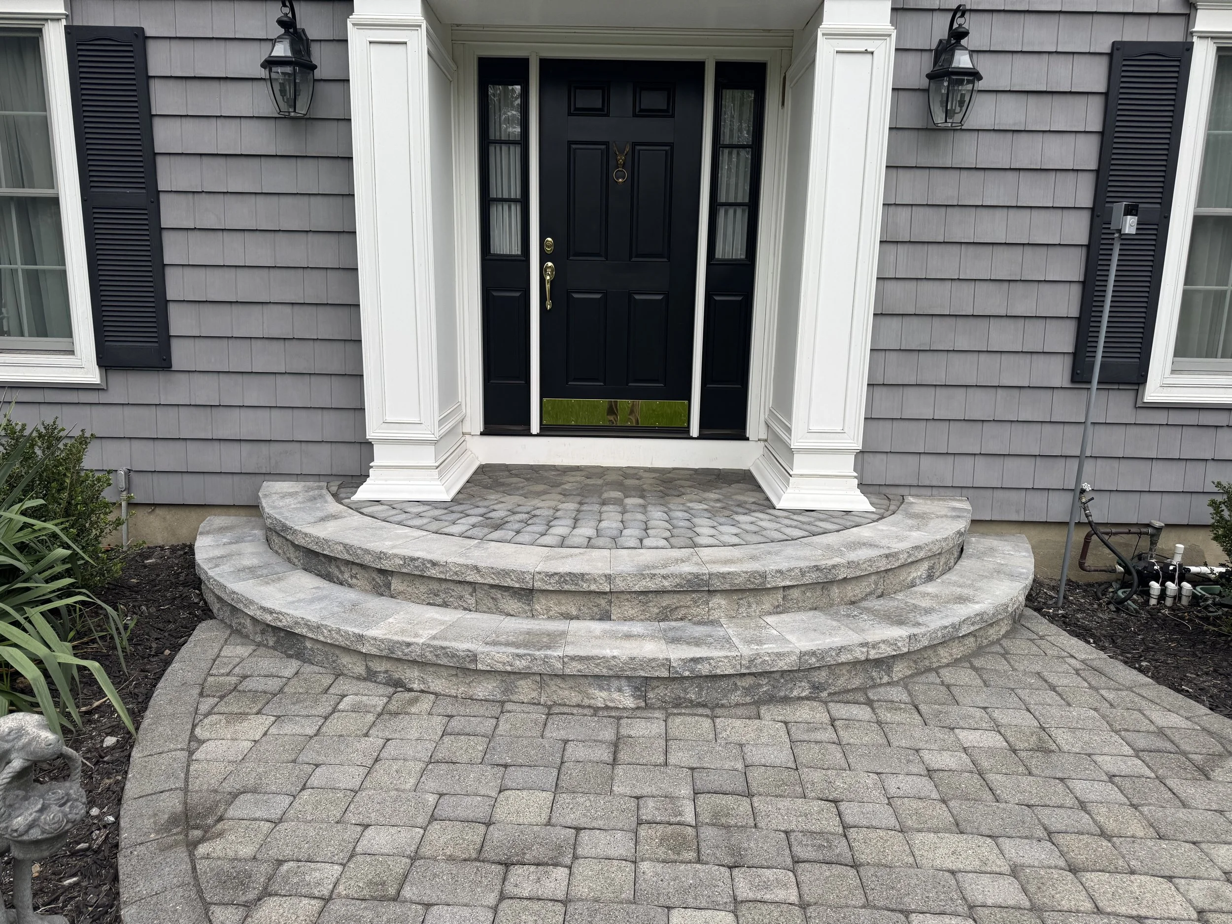 Front entrance of a house with a black door, gray shingle siding, two black shutters, and a decorative white columned porch with cobblestone steps.