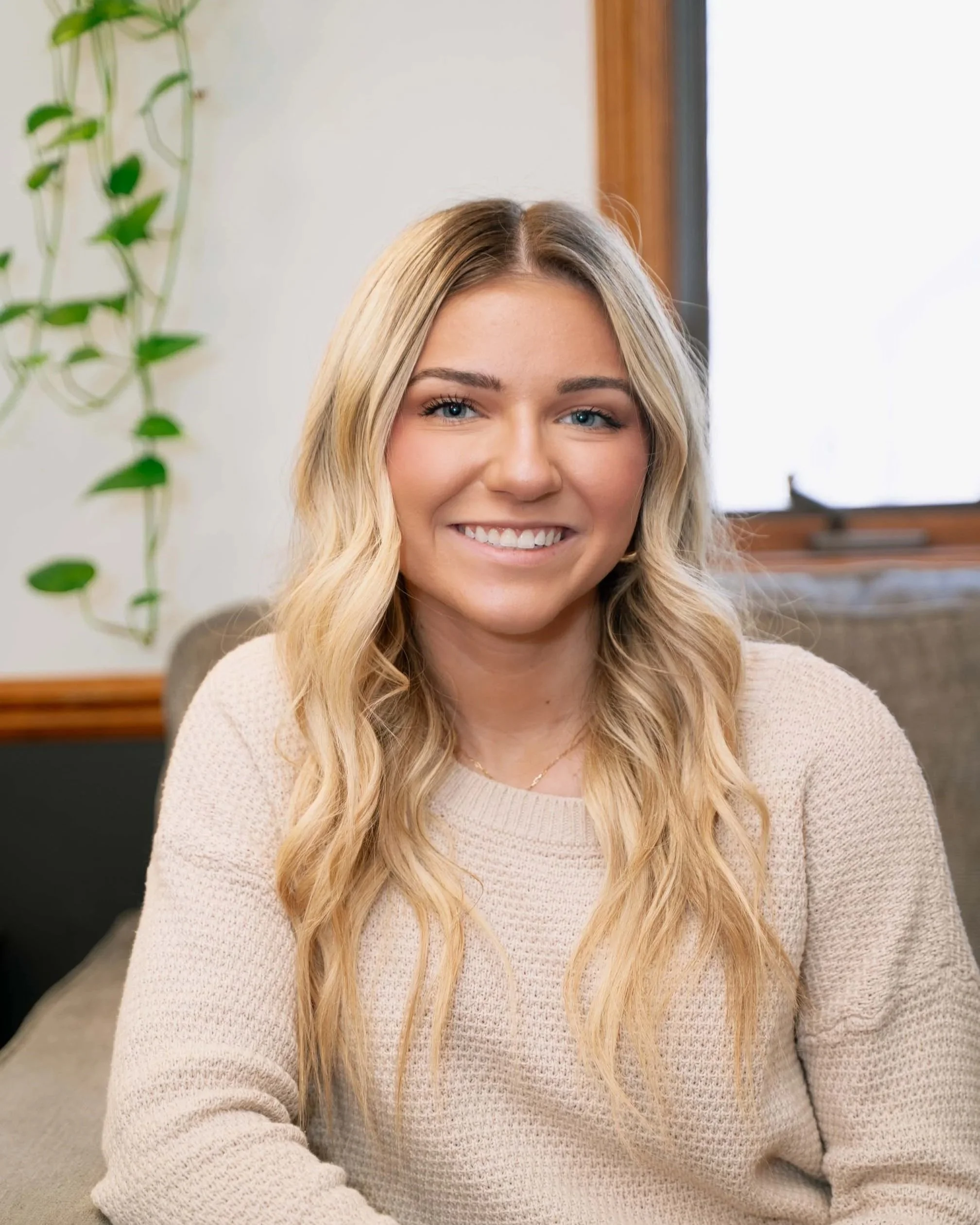 Smiling woman with long blonde wavy hair sitting on a beige couch in a room with a plant and a window in the background.