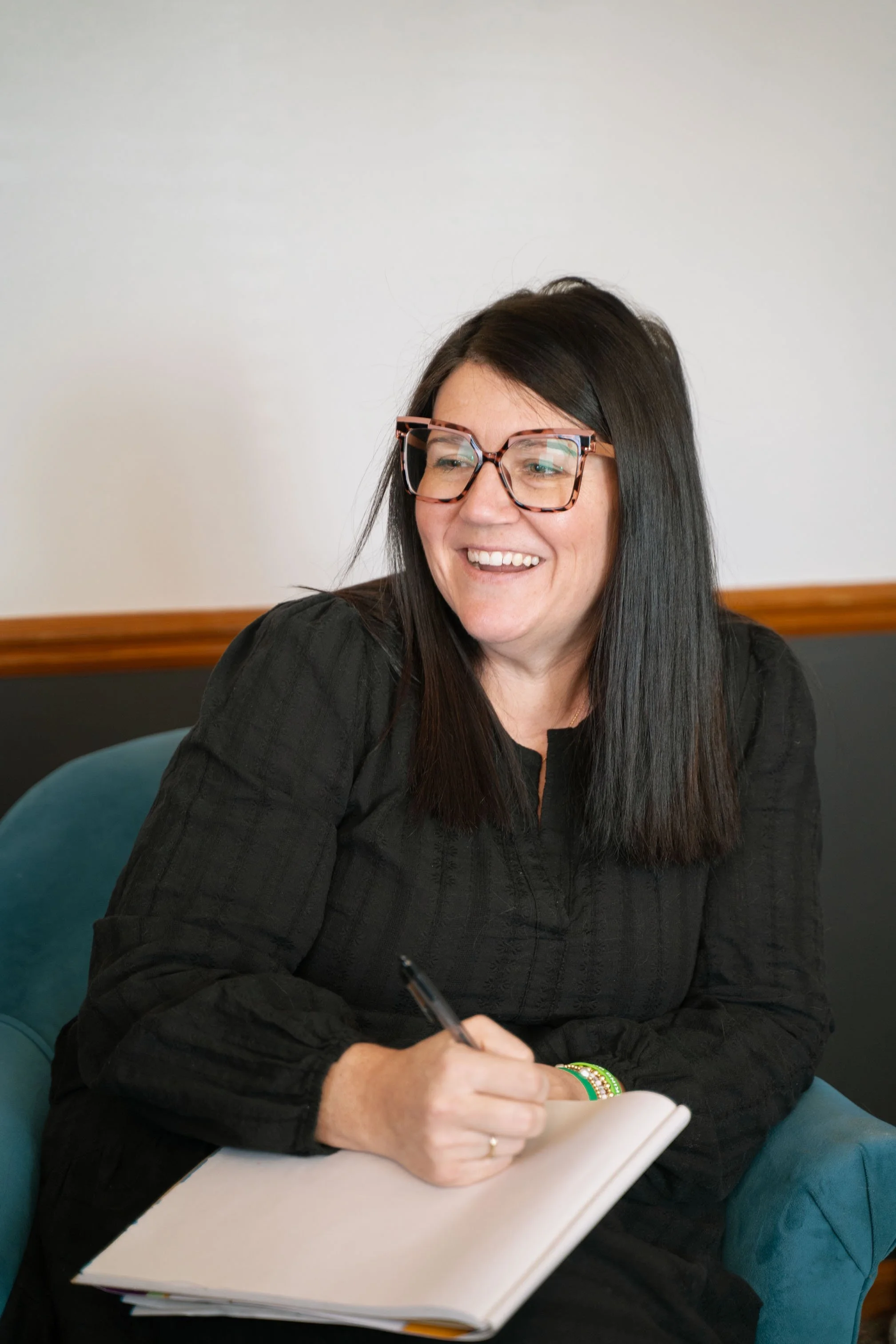 A woman with black hair, glasses, and a black shirt sitting on a teal chair, smiling and holding a notebook and pen.