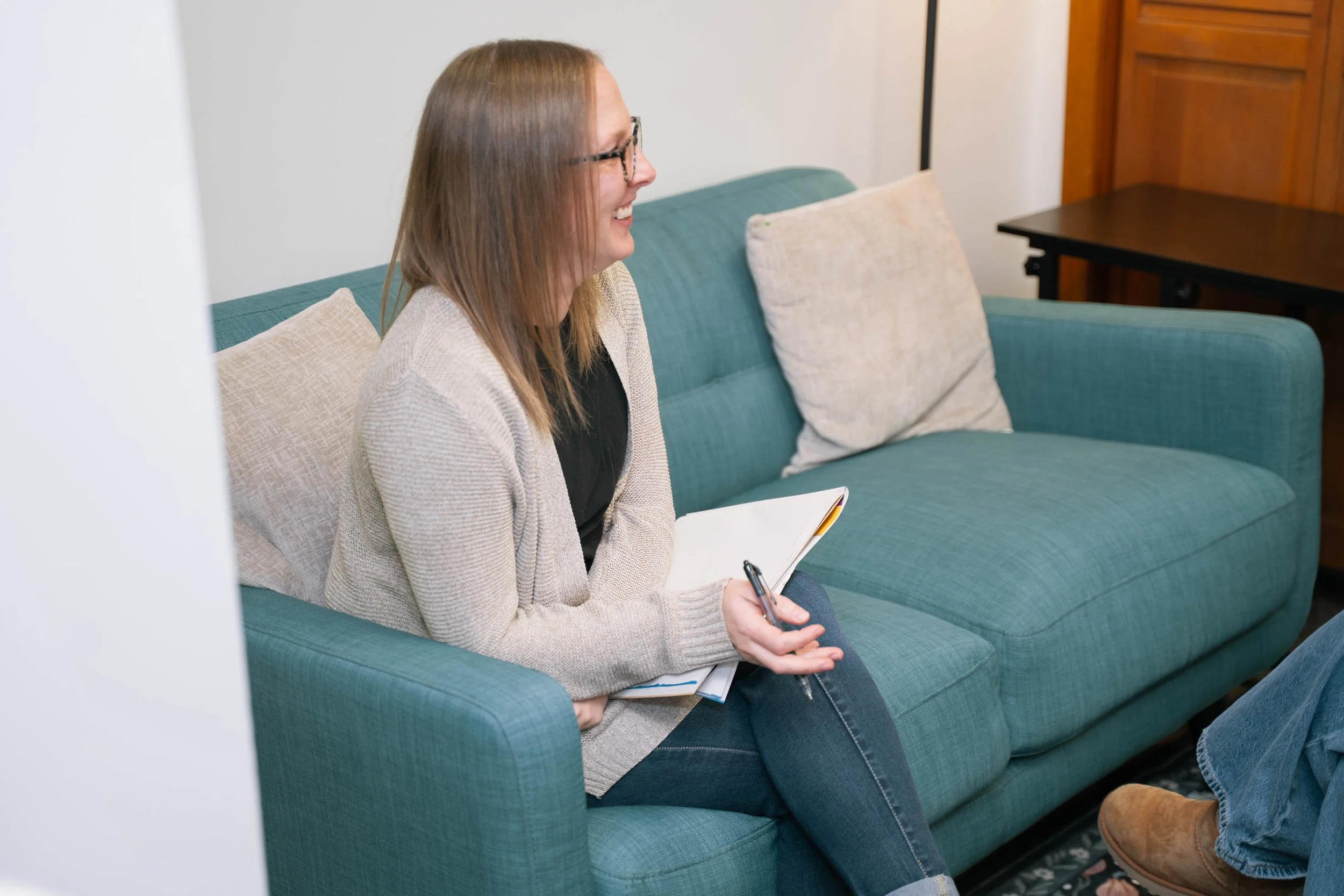 A woman with glasses and shoulder-length hair sitting on a teal sofa, smiling, holding a notebook and pen, wearing a beige sweater and black top, with a beige pillow behind her, in a cozy room with wooden furniture.