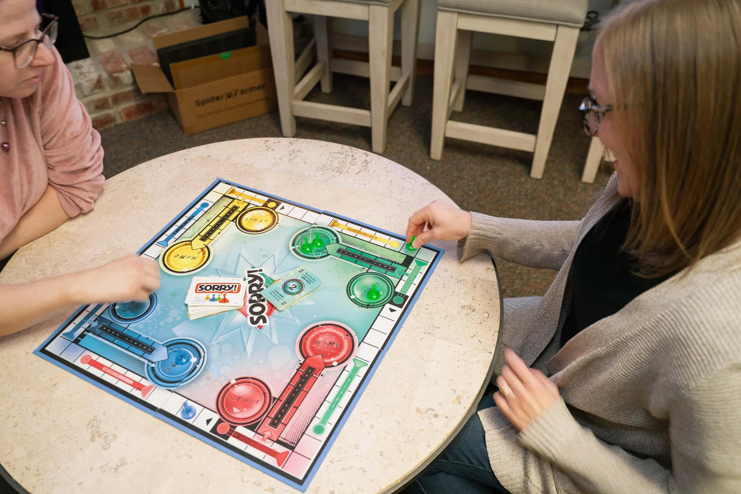 Two women playing Monopoly on a round table, with game pieces, cards, and 'Sorry' cards visible.