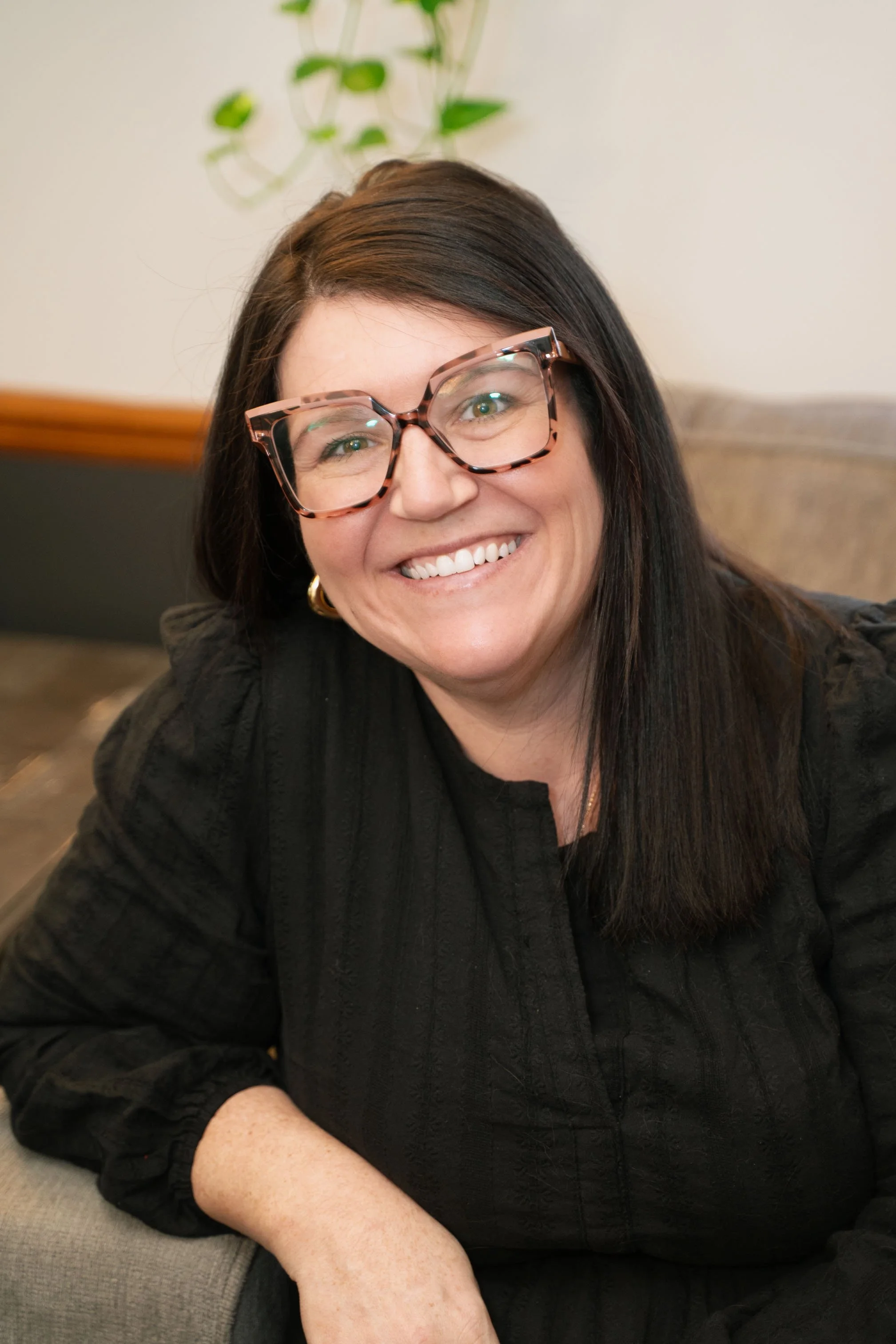 A woman with long dark hair and glasses smiling while sitting indoors.