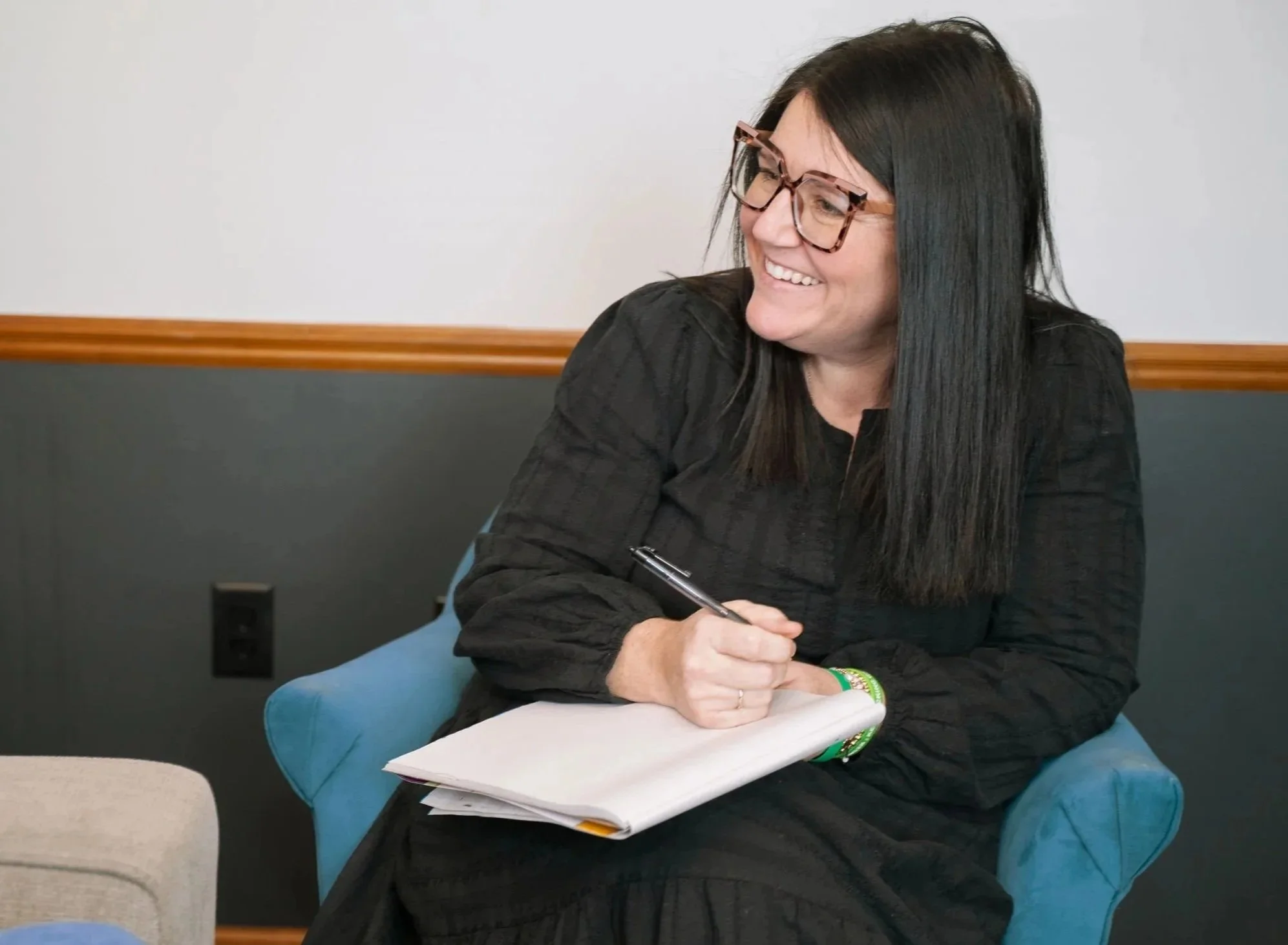 A woman with long black hair, wearing glasses and a black dress, sitting on a blue chair, smiling, holding a pen and a notebook in a room with plain walls.