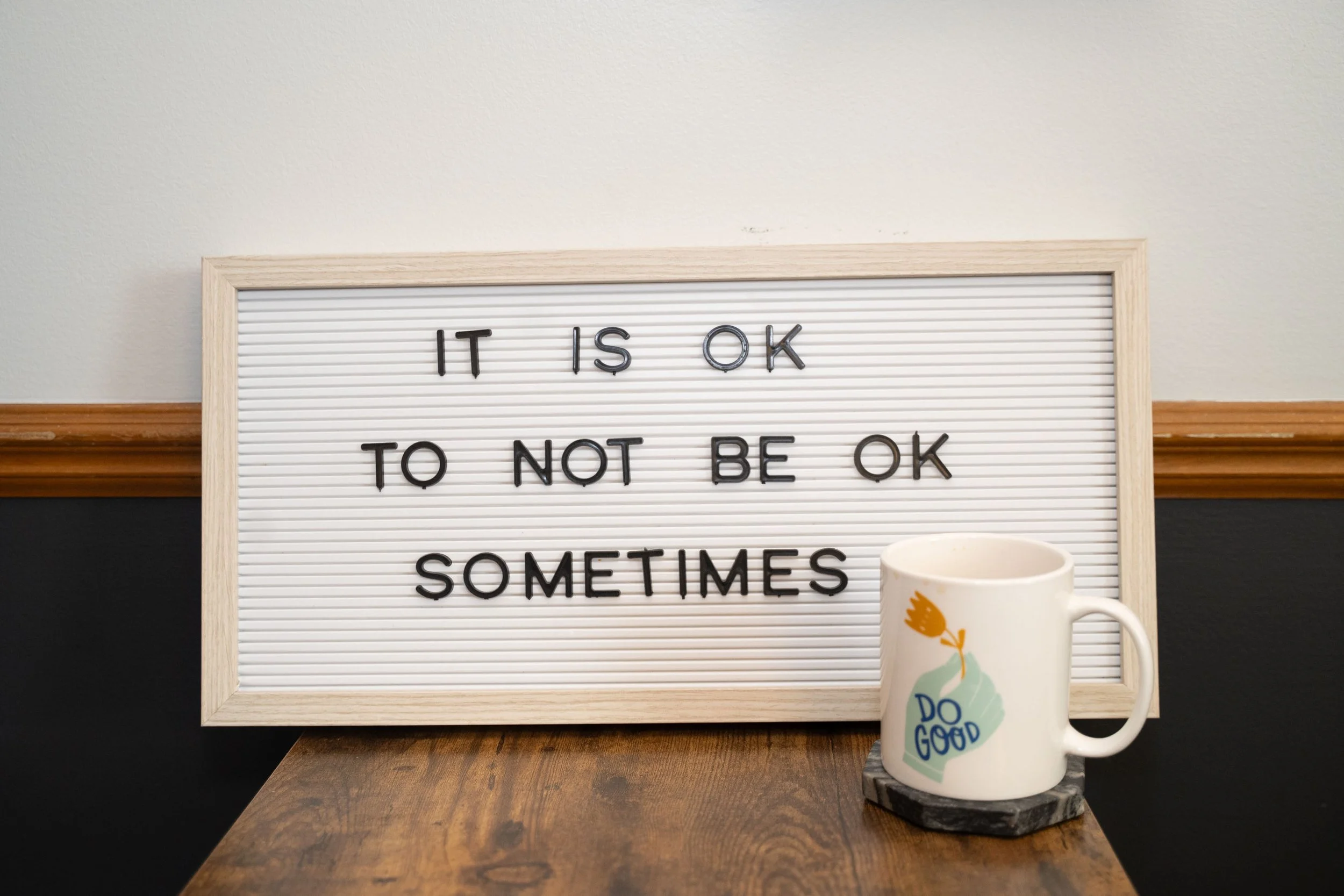 A whiteboard with the message 'It is ok to not be ok sometimes' and a mug with a 'Do Good' message on it sitting on a wooden table.