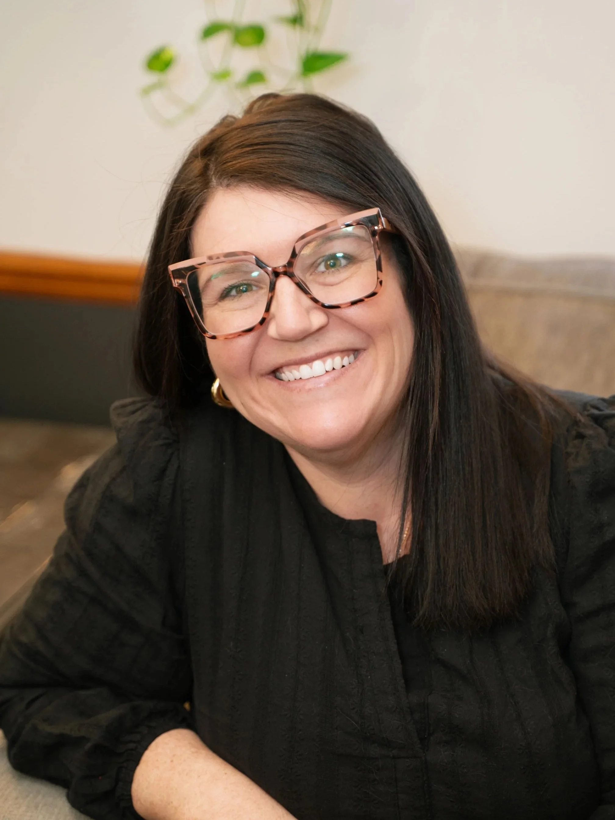 A woman with dark hair, wearing large pink tortoiseshell glasses, smiling at the camera, dressed in a black top, sitting in an indoor setting with a beige wall and a houseplant in the background.