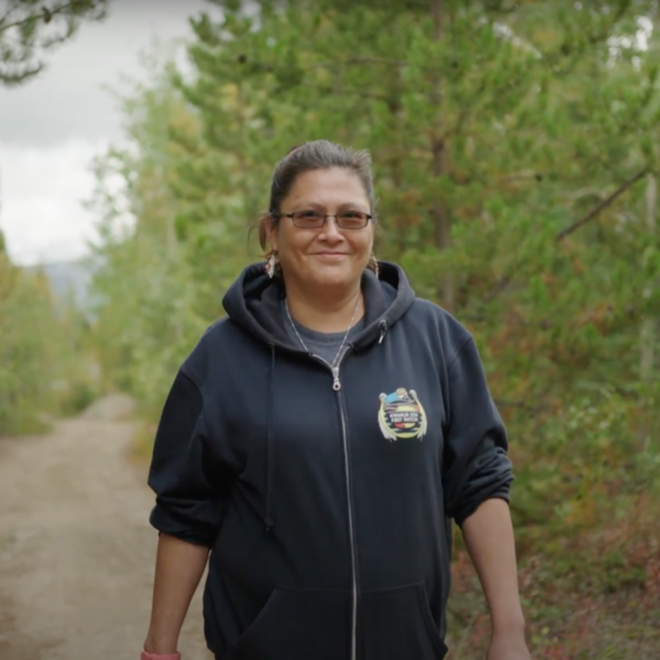 Woman with glasses and dark hair, smiling, wearing a black hoodie walking outdoors on a dirt path surrounded by green trees.