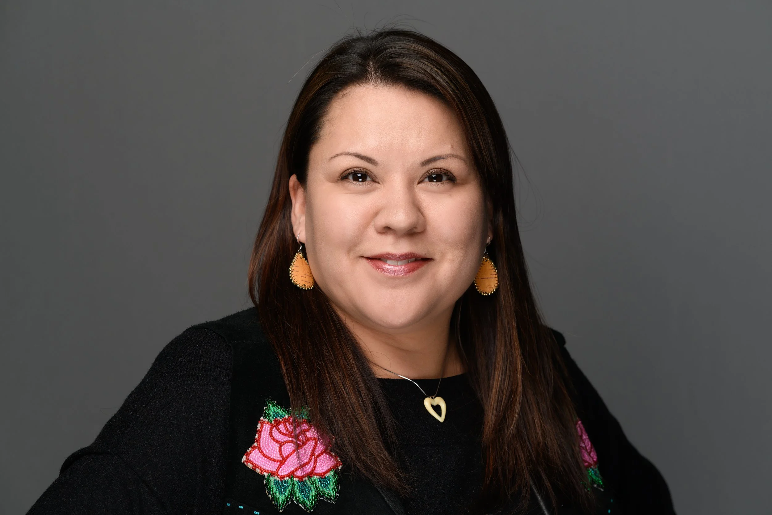 Headshot of a woman with long brown hair, smiling, wearing gold and orange earrings, a necklace with a heart-shaped pendant, and a black top with colorful embroidered flowers on the shoulder, against a gray background.