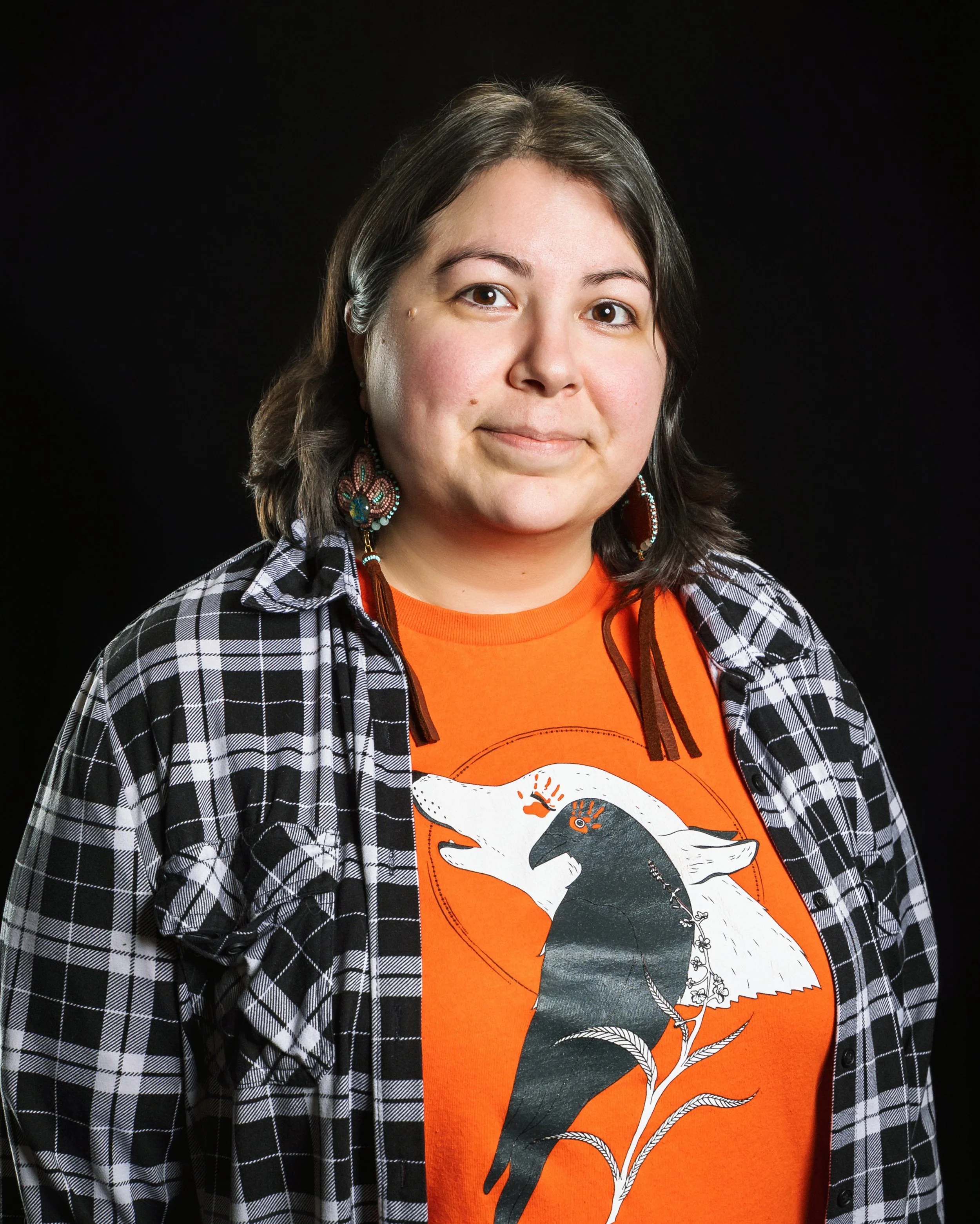 Portrait of a woman with shoulder-length dark hair, wearing a black and white checkered shirt over an orange T-shirt with a black and white bird illustration, and colorful earrings, against a black background.