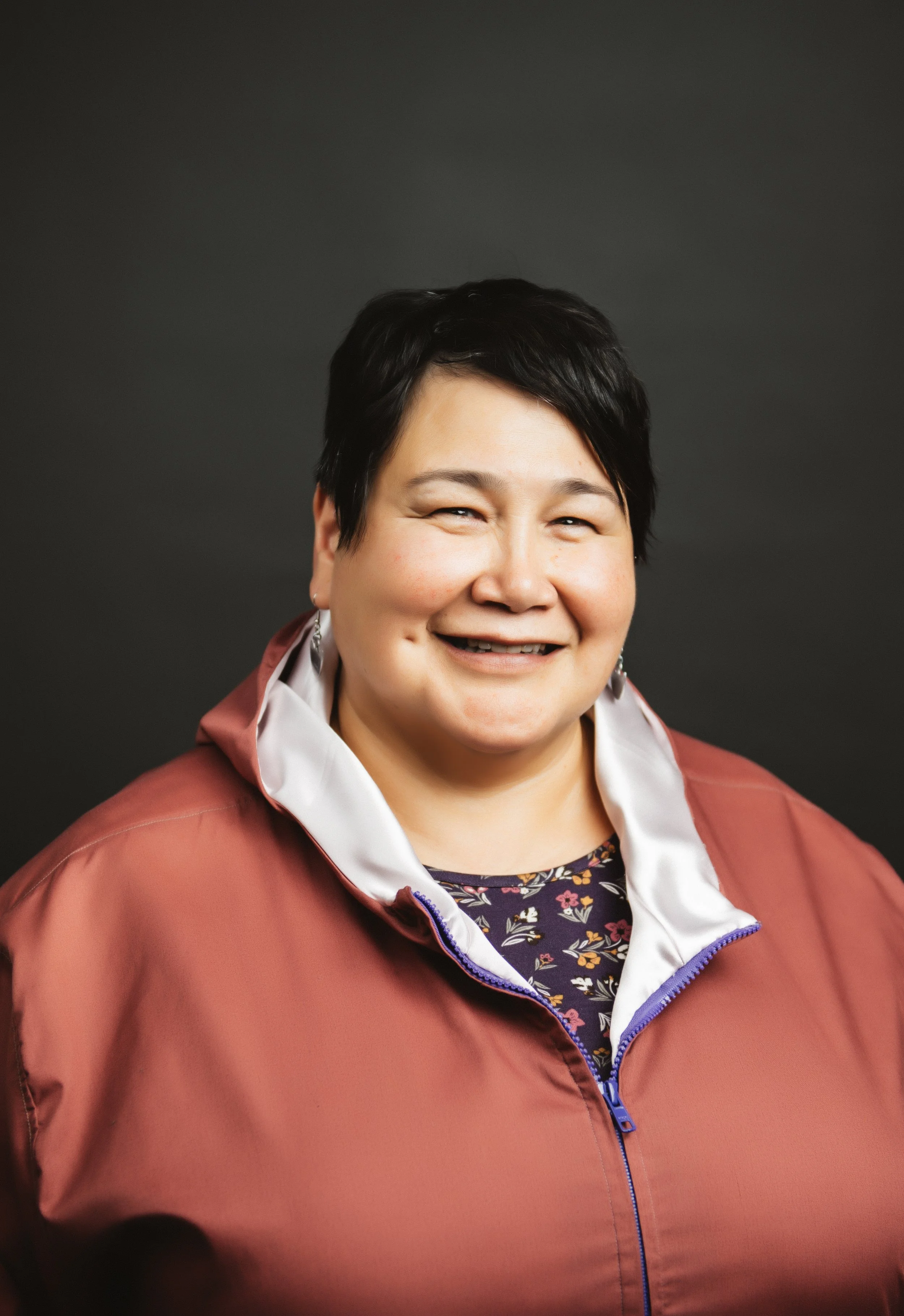 A smiling woman with short black hair, wearing a red jacket with a white collar and a floral top, poses against a dark background.