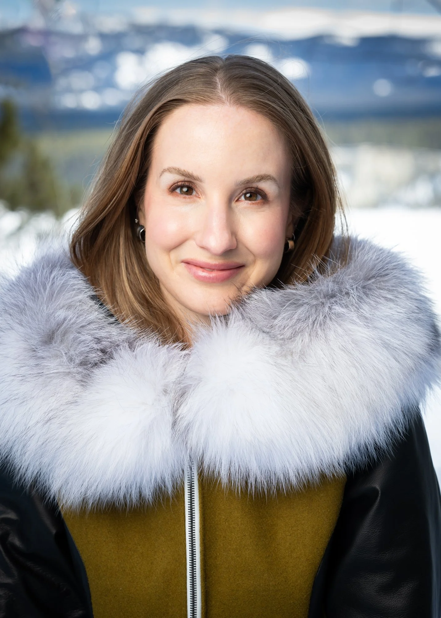 Close-up portrait of a woman with light brown hair, smiling, wearing a jacket with a large fur collar, outdoors with blurred winter landscape background.