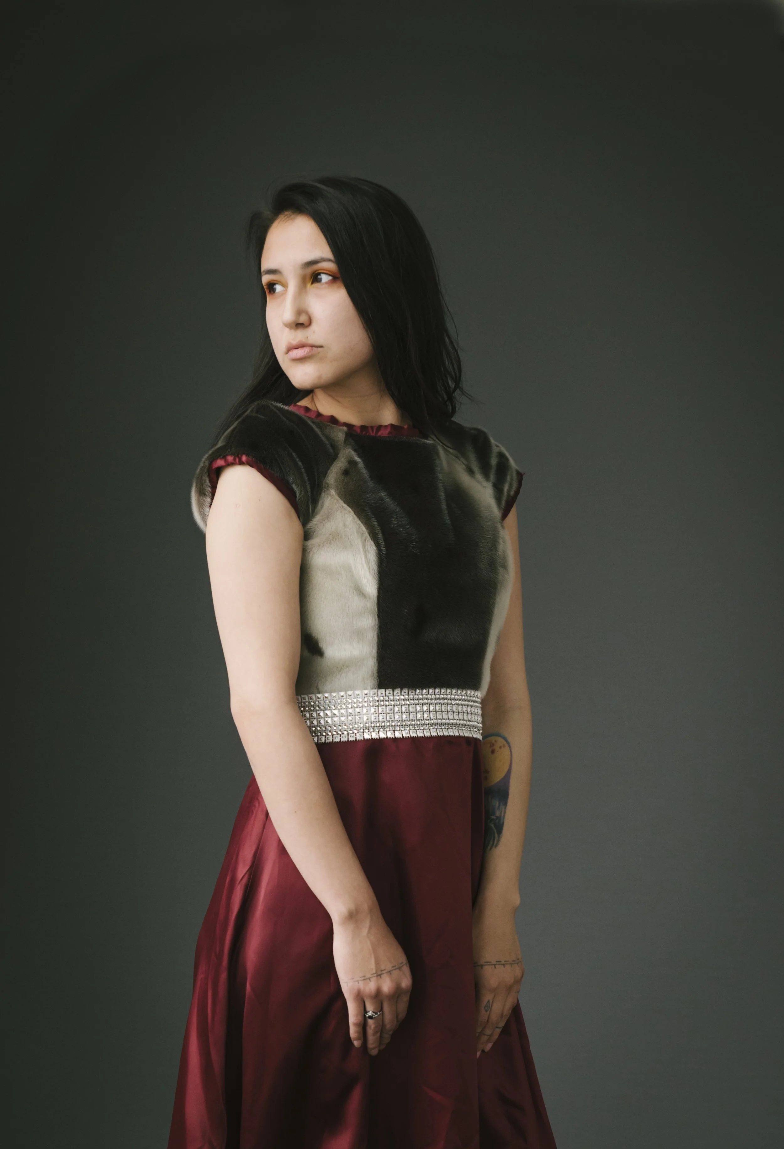 Young woman with black hair wearing a black and silver top and a long maroon skirt standing against a dark background.