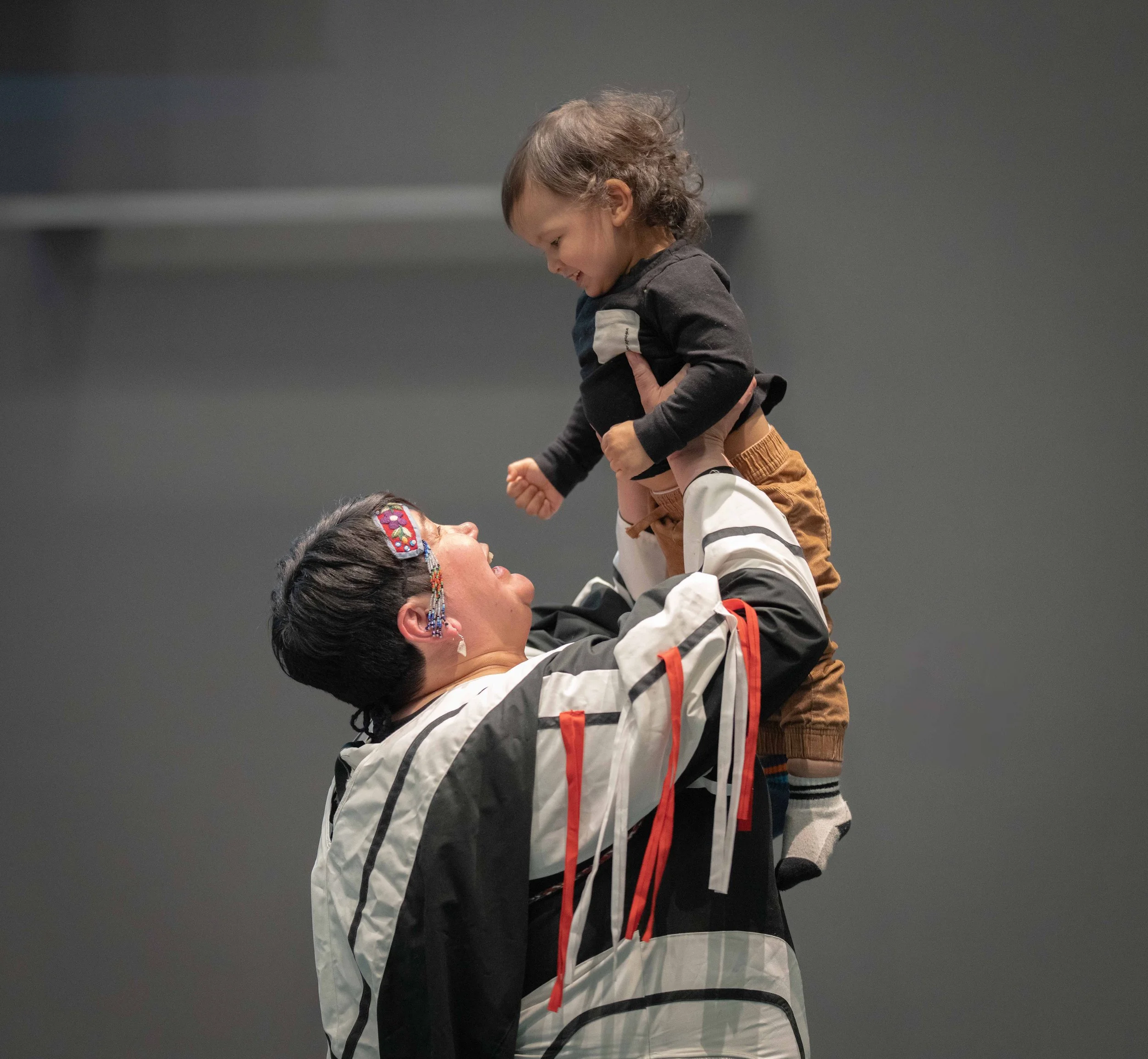 A woman with colorful beads and glasses lifting a young child in the air, both smiling and looking at each other.