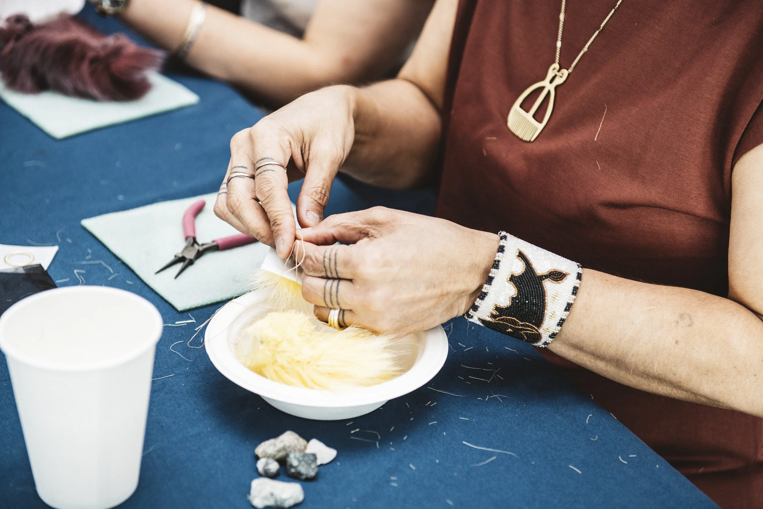Person working on jewelry making, handling a small piece with needle and thread, surrounded by tools on a blue table.