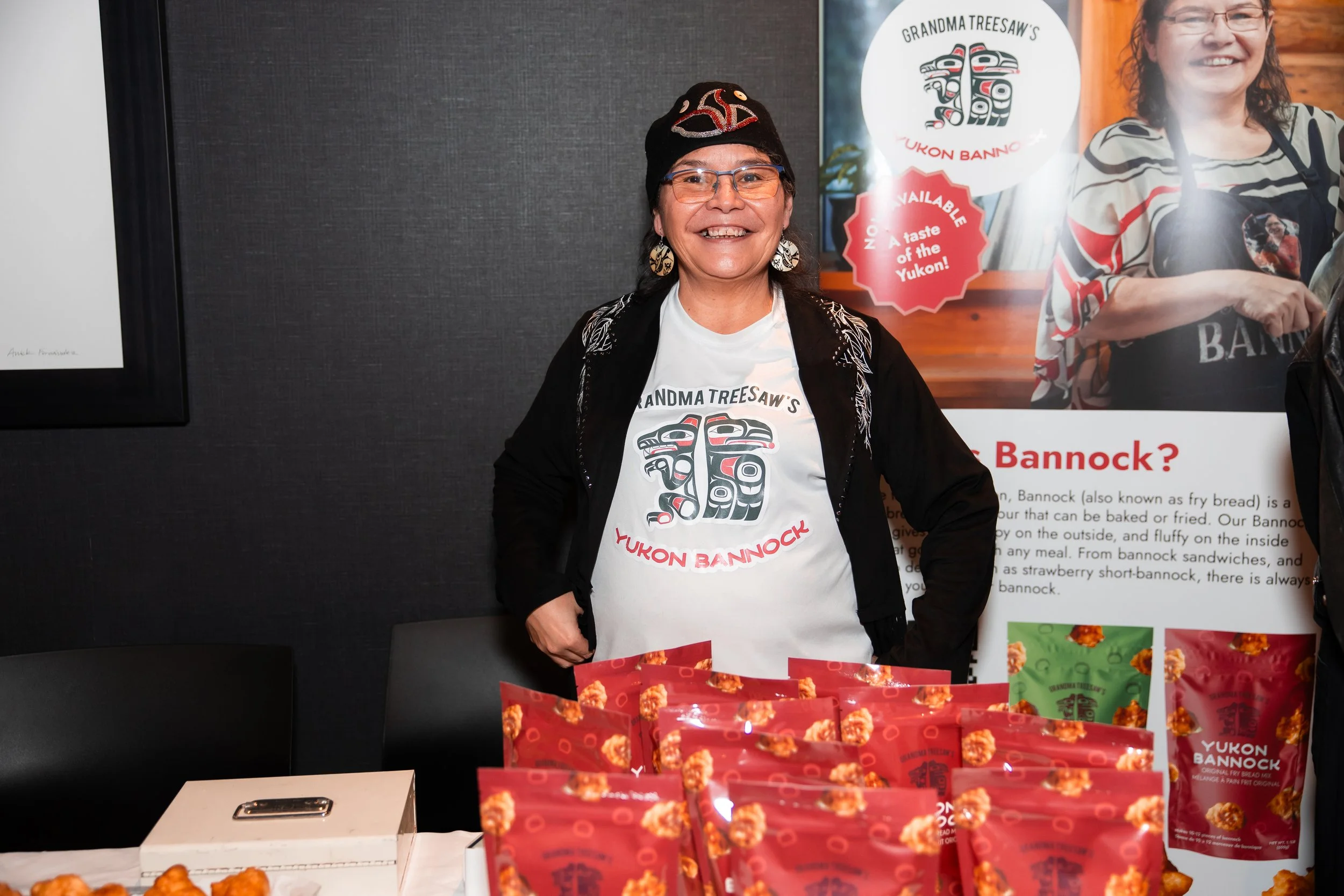 Smiling woman wearing glasses and a black hat standing behind a table with red packages of Yukon Bannock, in front of a promotional poster about Bannock.